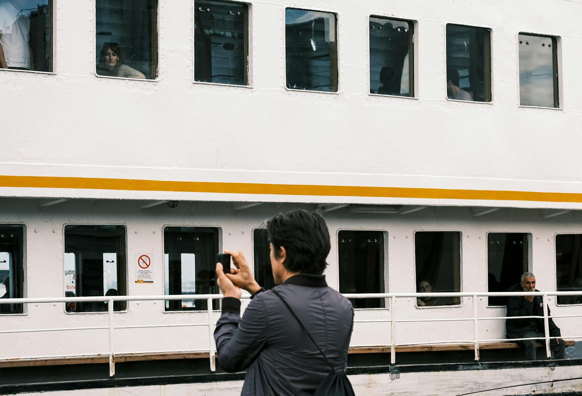 Man takes photo of ferry with passengers visible through windows at daytime.