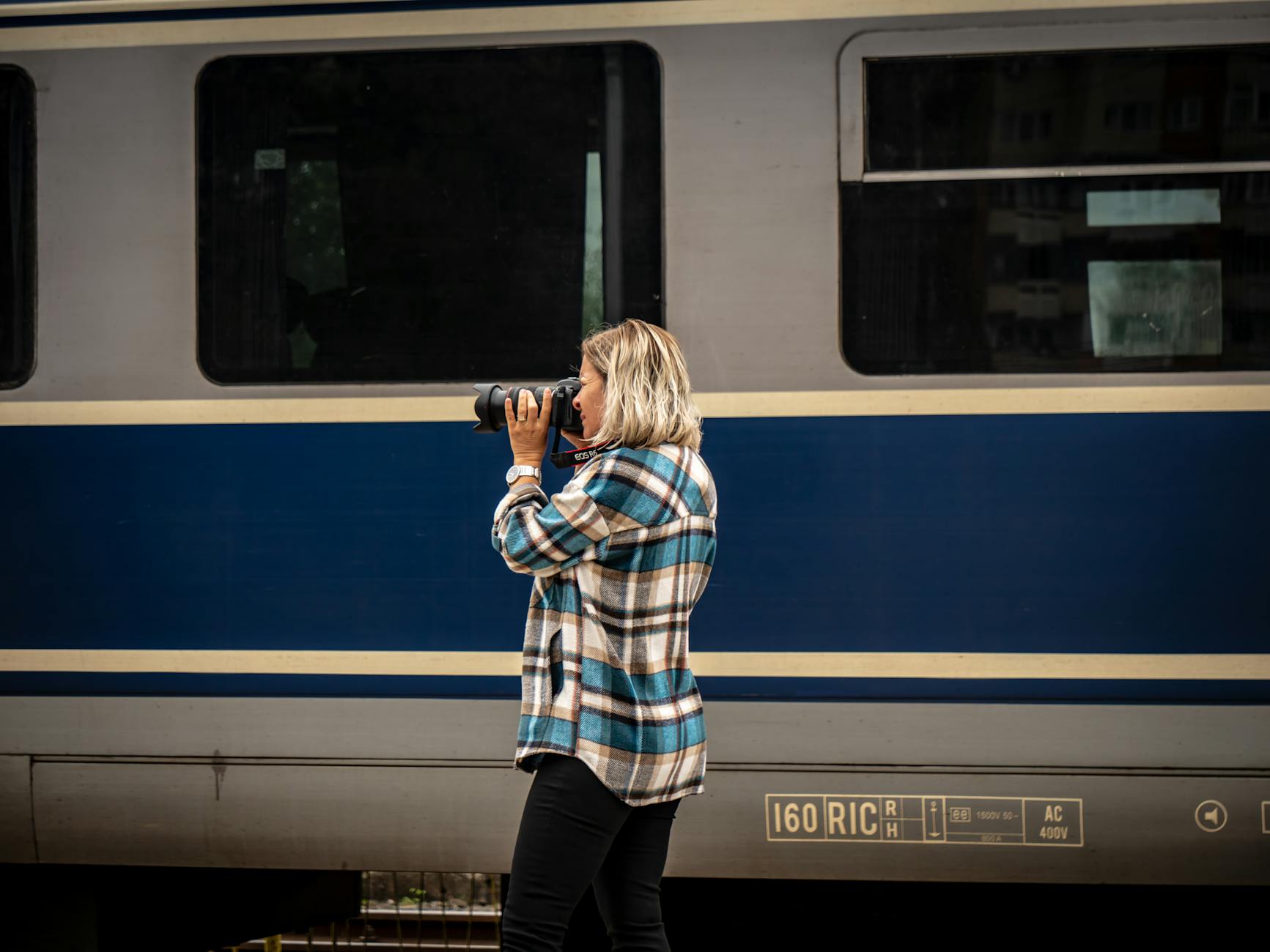 A woman in a plaid shirt capturing a train with her camera outdoors.