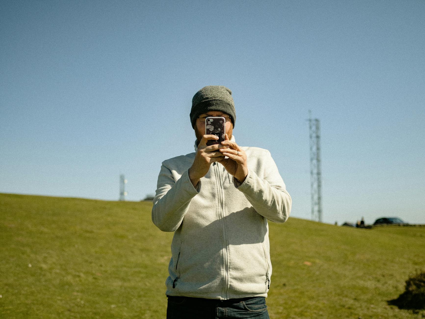 Concentrated male in hat taking picture on mobile phone while standing on lush grassy meadow and looking at camera