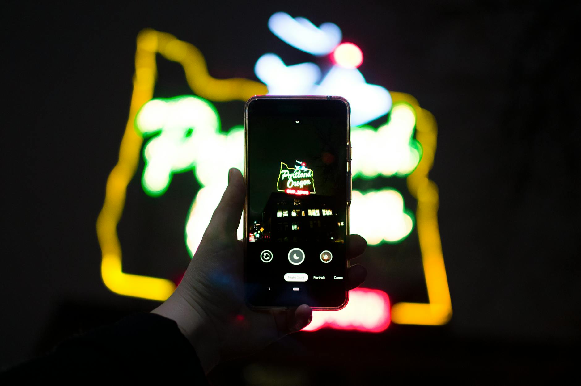 Hand holding smartphone capturing neon 'Portland Oregon' sign during night in downtown Portland.