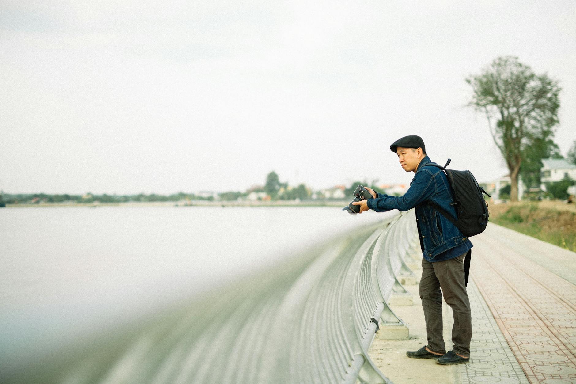 Photographer stands by riverside rail capturing the serenity of a cloudy day. Artistic and calm.
