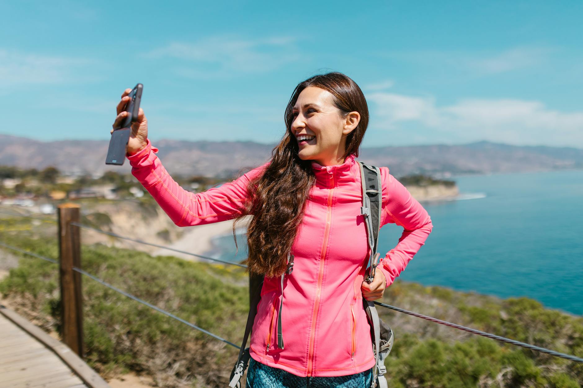 Happy woman in pink jacket takes a selfie on a coastal hiking trail.