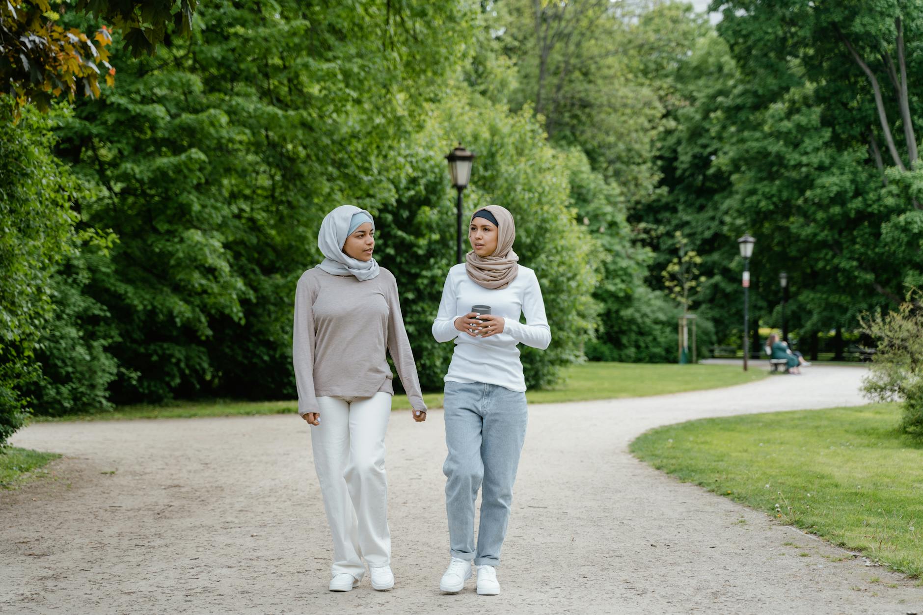 Two women in hijabs walking and conversing in a lush green park on a sunny day.