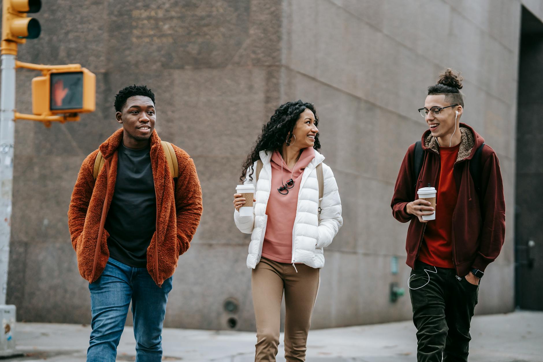 Positive young diverse male and female teens smiling and chatting while walking on city street with takeaway coffee in hands