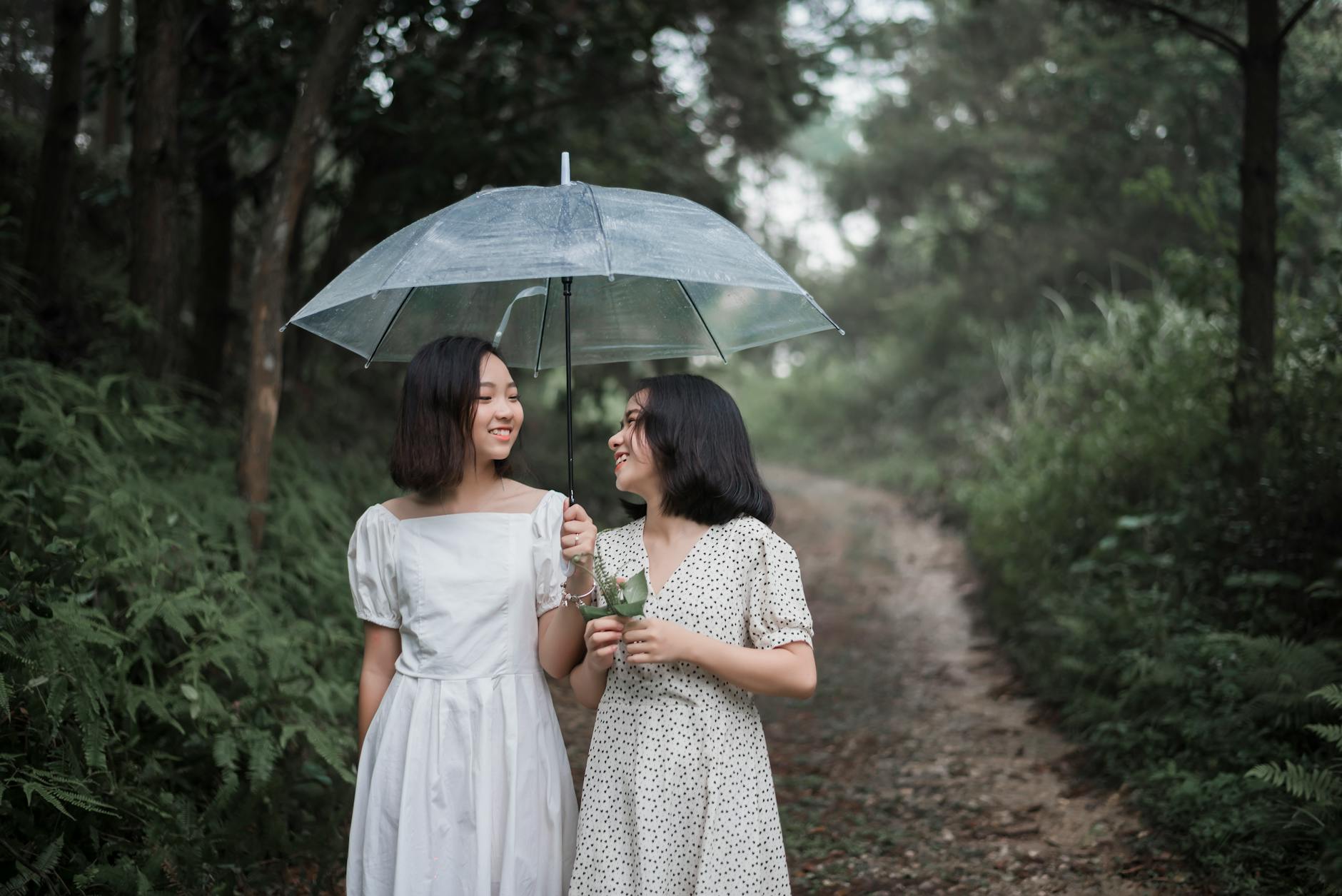 Two young women share a joyful walk under an umbrella through a lush forest trail.