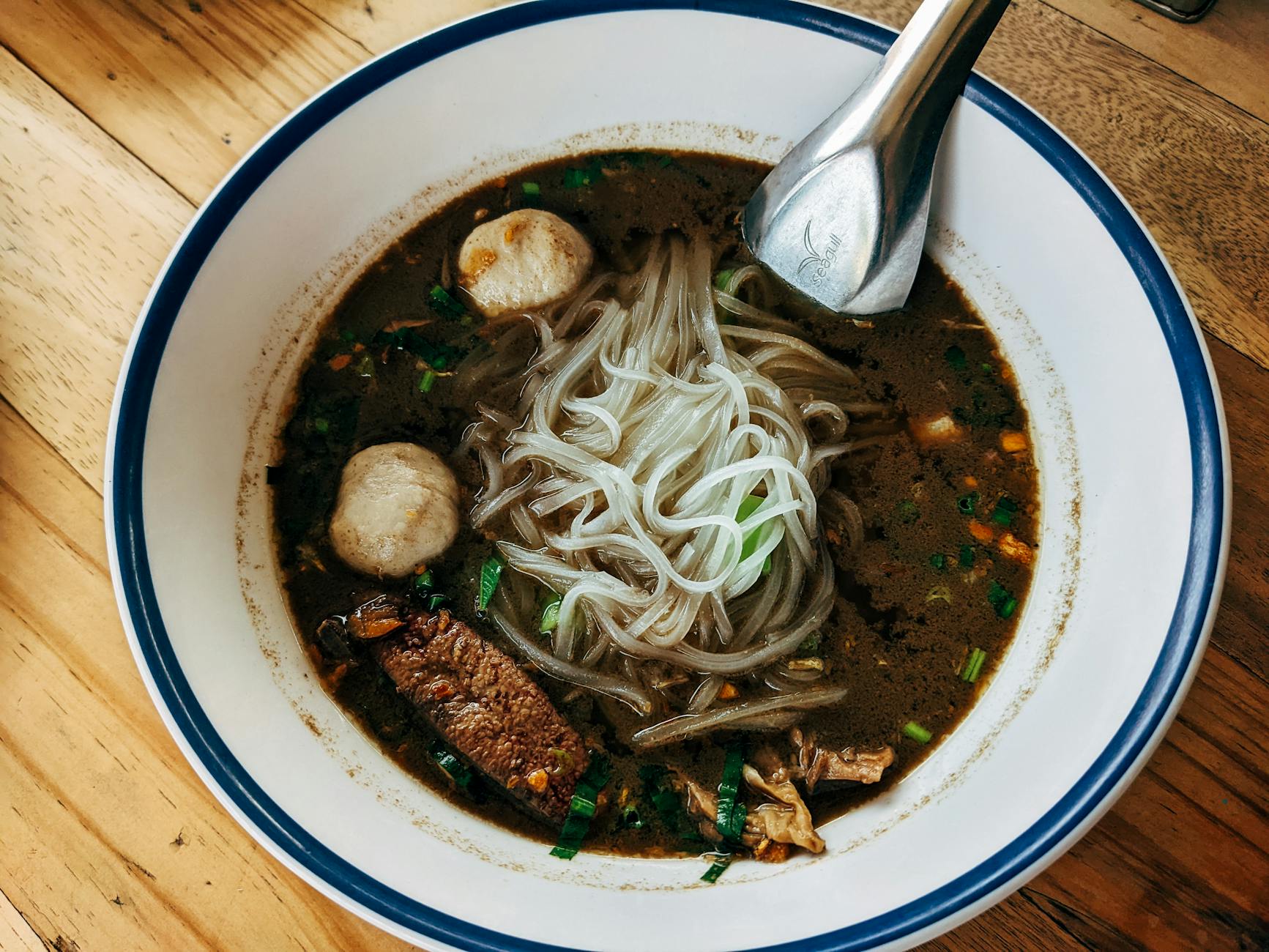 High angle shot of flavorful Thai beef noodle soup in a ceramic bowl on wooden surface.