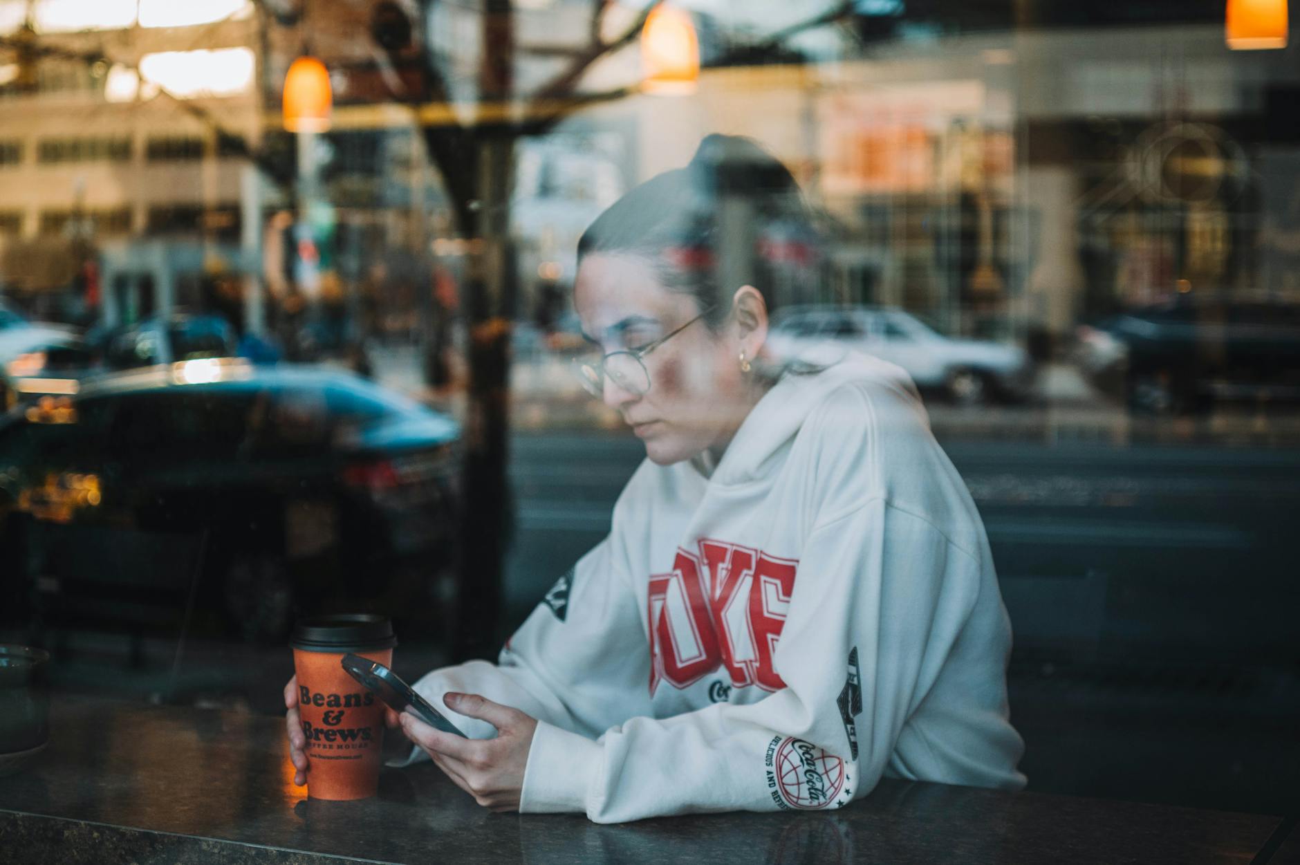 A woman in a hoodie sits inside a cafe, holding a disposable cup and mobile phone, seen through glass.