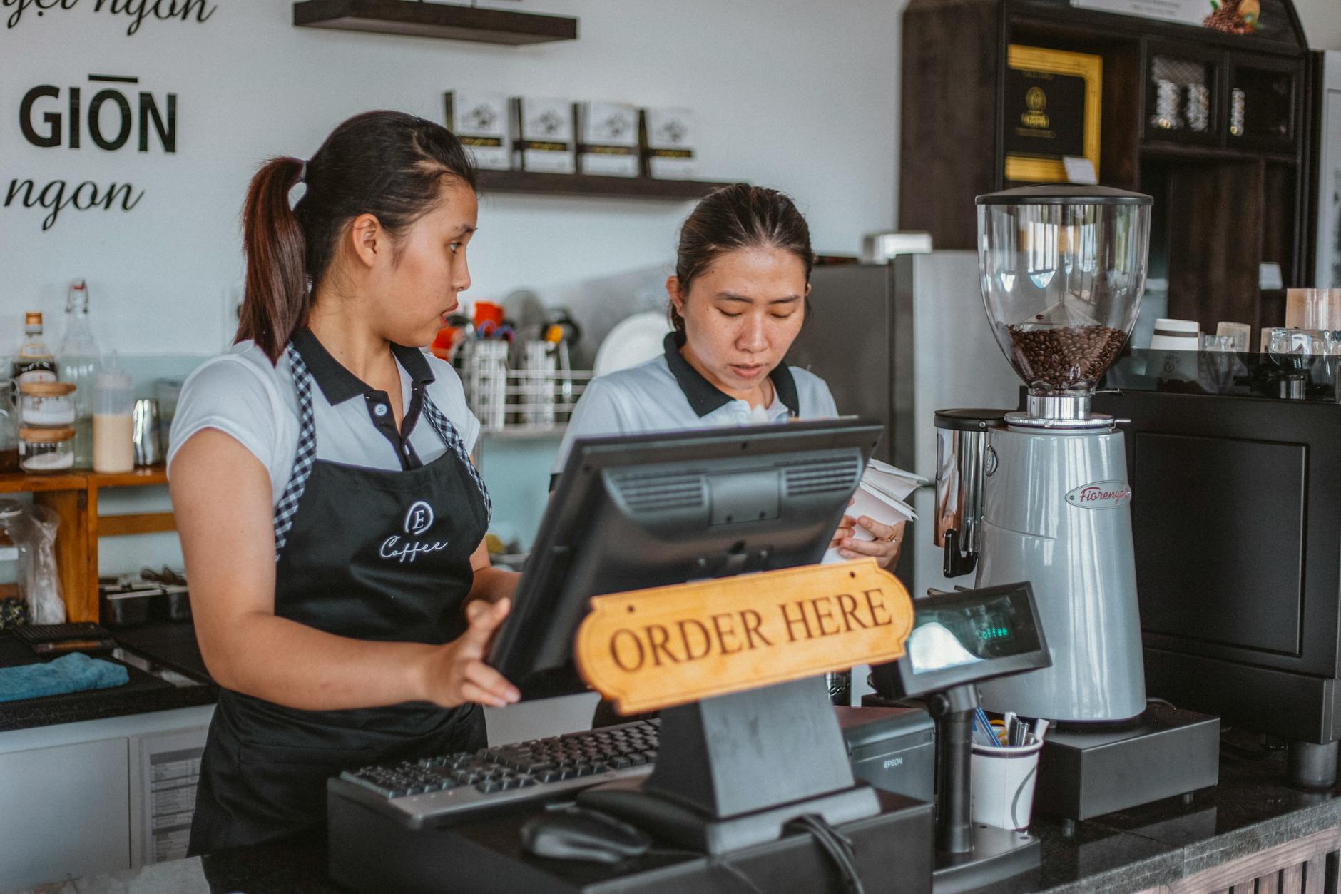 Two baristas work at a coffee shop counter with a cash register and equipment.