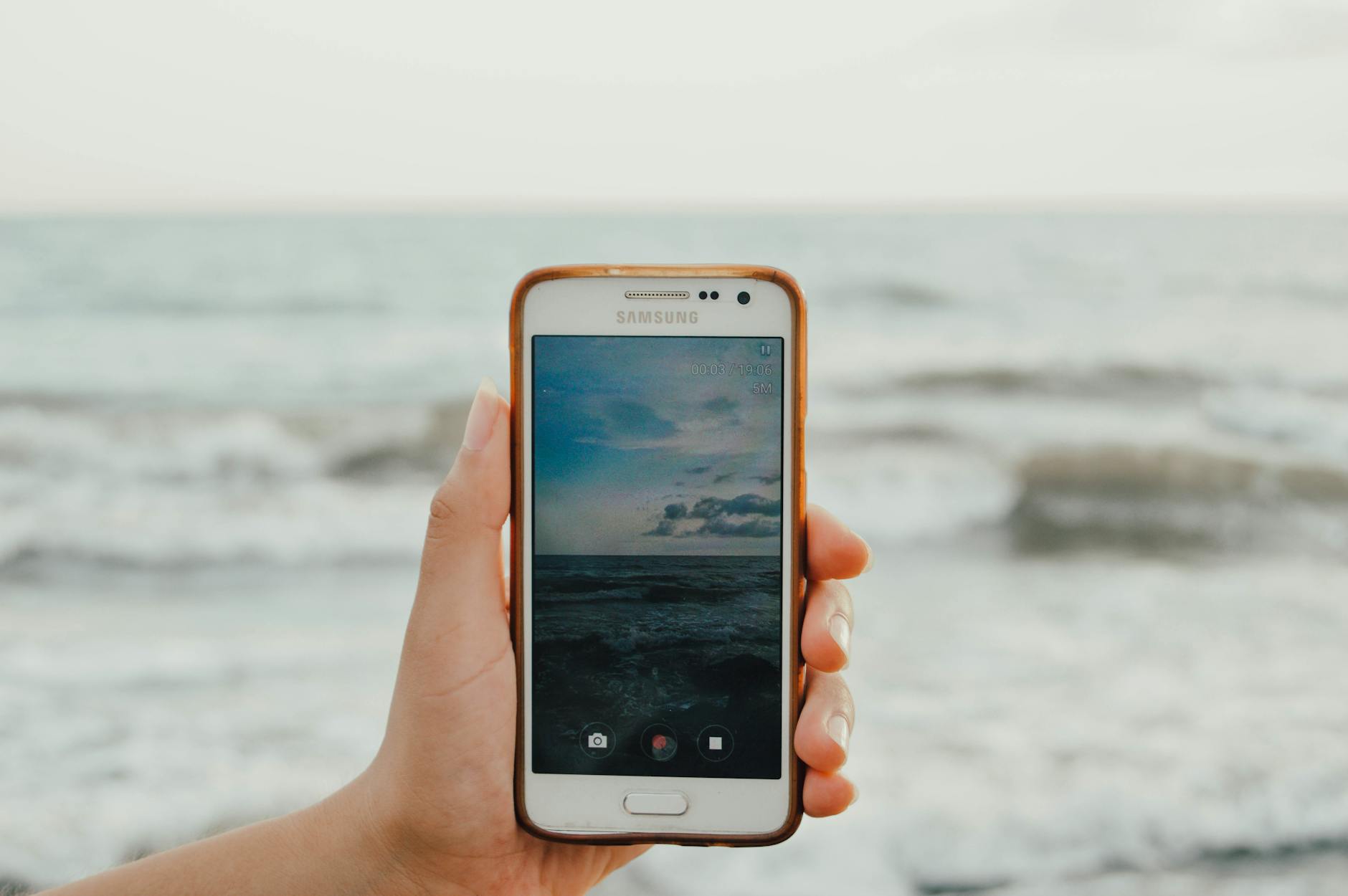 A hand holds a smartphone capturing a picturesque view of the ocean waves at the beach.