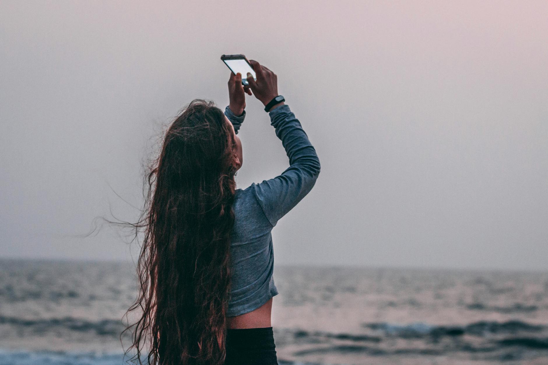 A young woman takes a photo of the sunset over the ocean, embracing leisure and travel vibes.