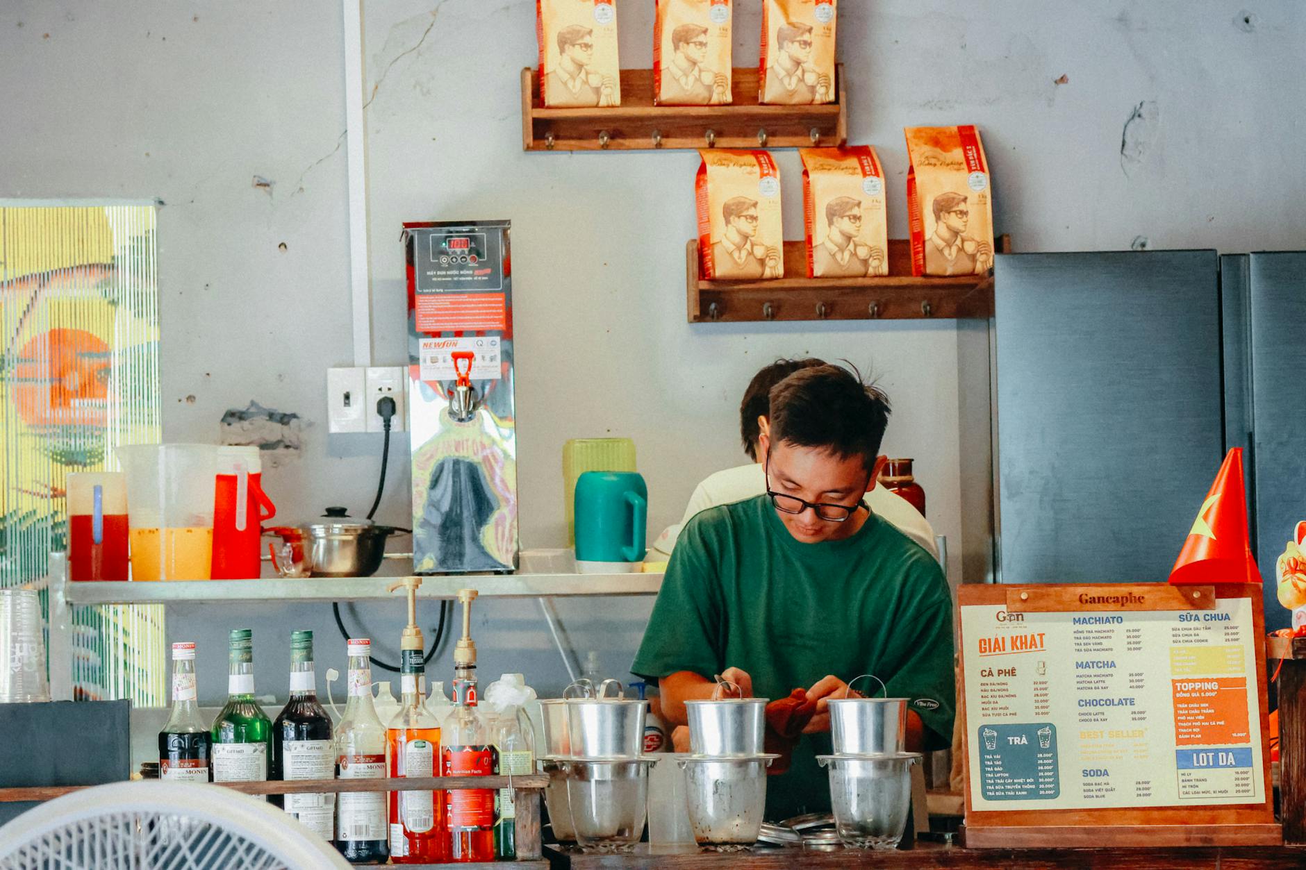 A barista making coffee in a Vietnamese cafe with various drinks and decor.