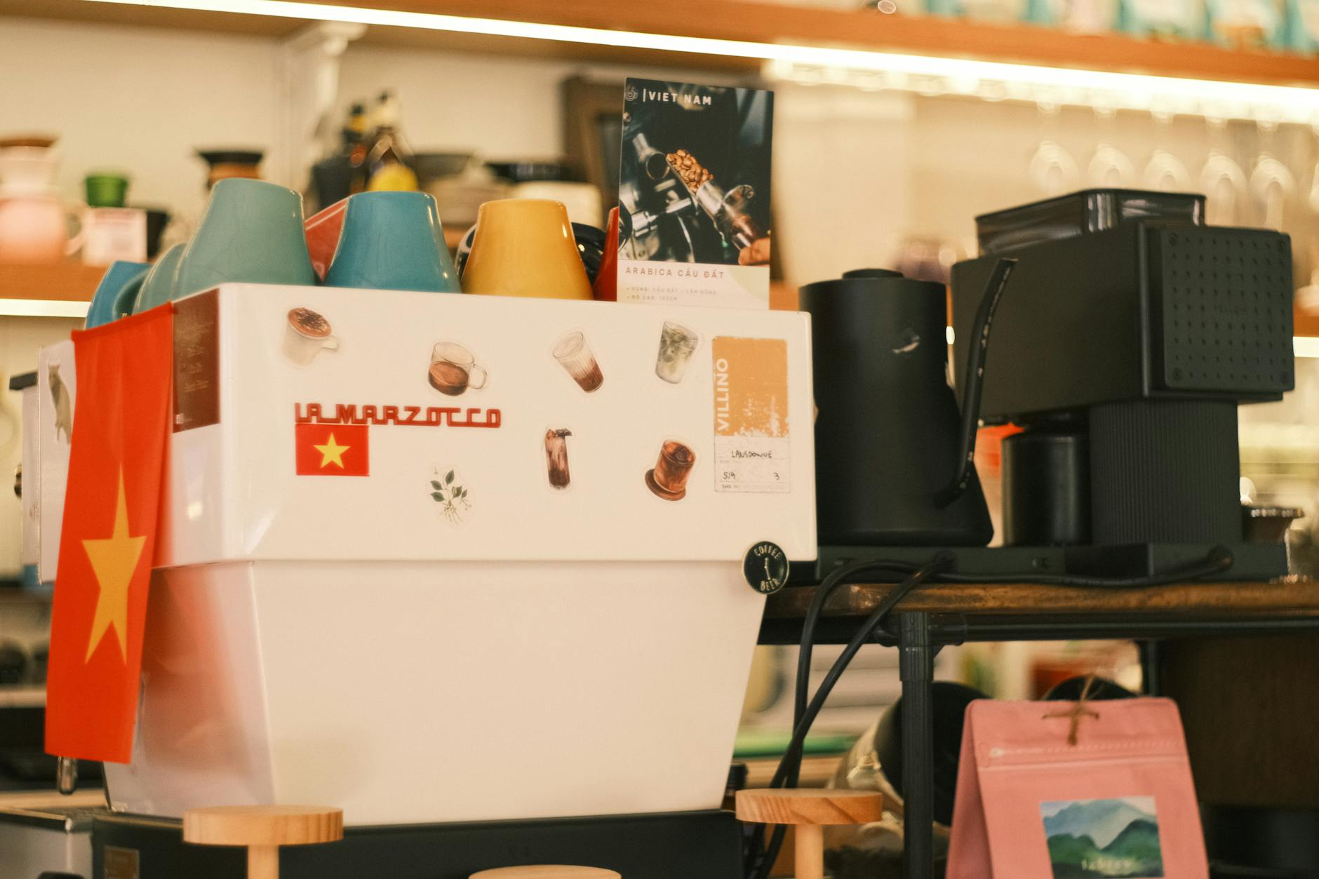 A cozy coffee shop with a La Marzocco espresso machine adorned with colorful cups and a Vietnam flag.