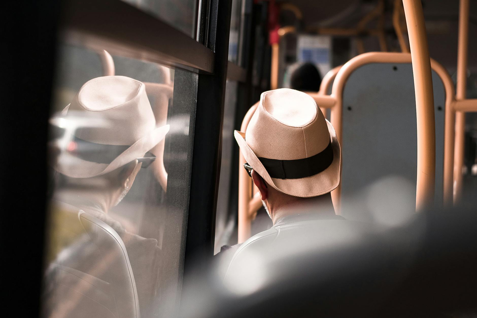 A man wearing a fedora hat sits inside a bus, reflecting on the window during a daytime commute.