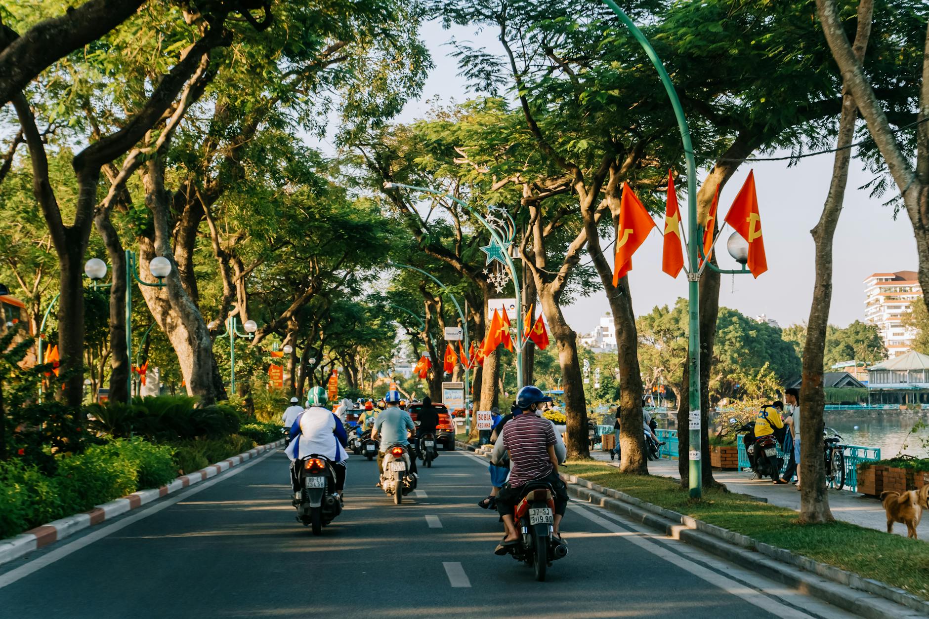 Motorbikes ride along a tree-lined street with flags in Hanoi, Vietnam during sunset.