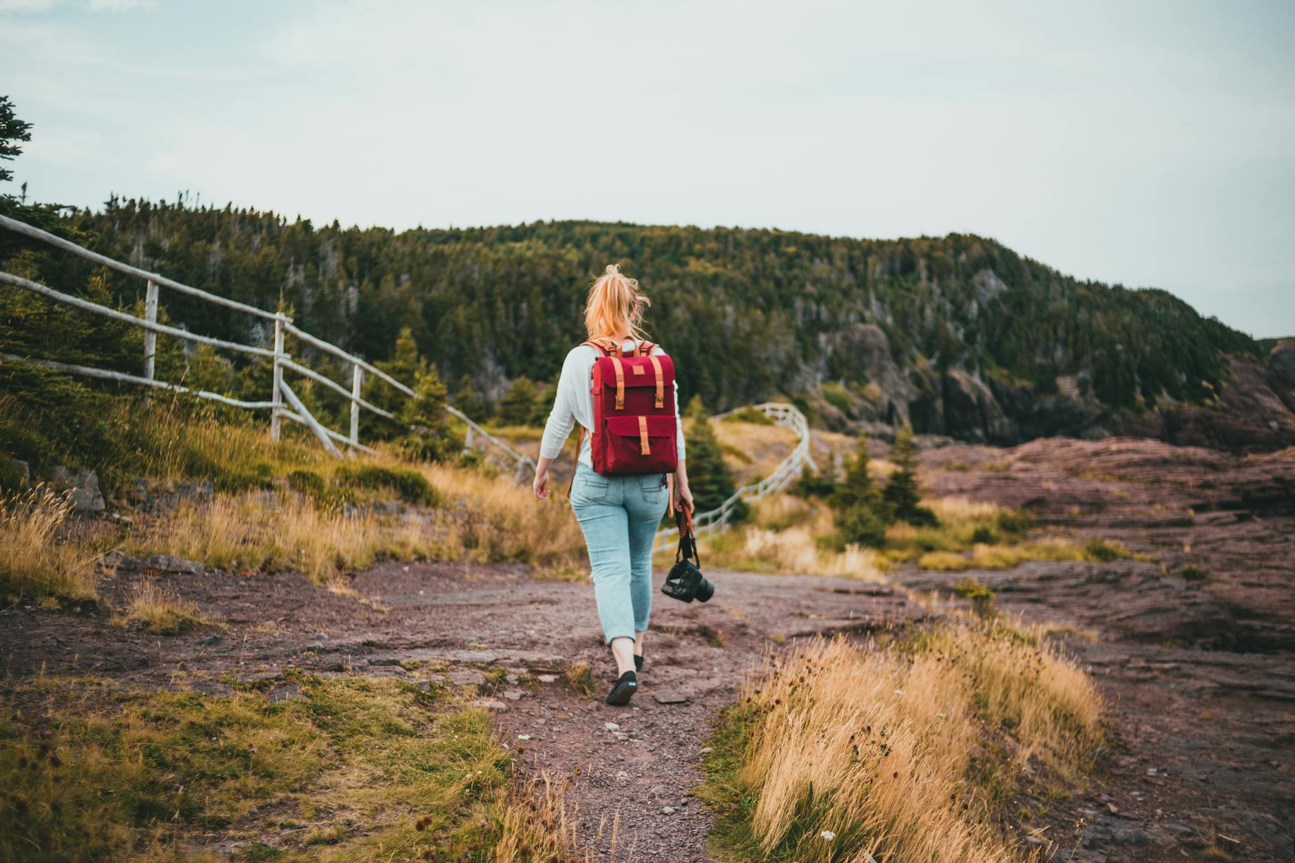 A young woman with a red backpack walks along a scenic path holding a camera.