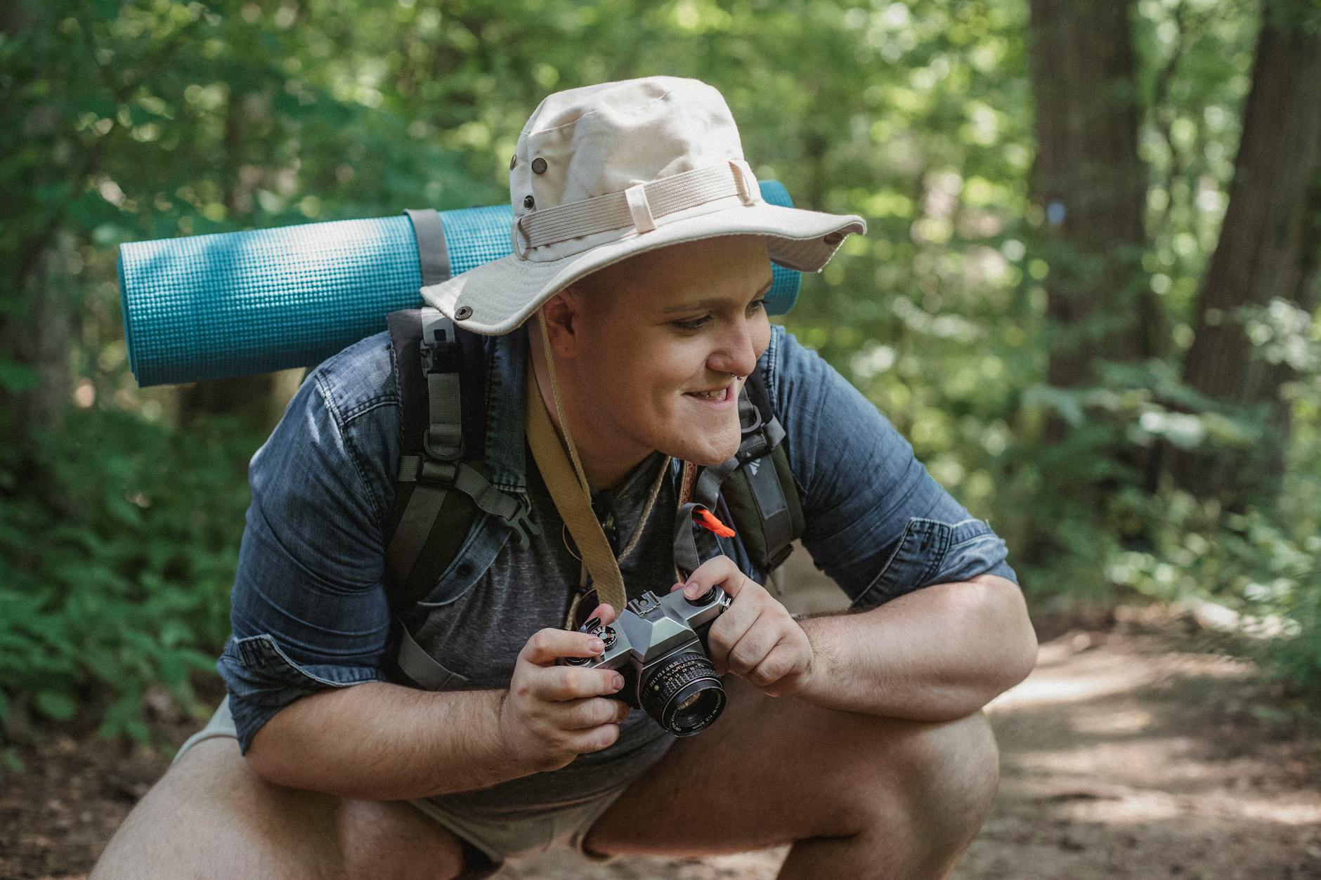 Young smiling male hiker with mat and photo camera squatting on pathway with trees behind while looking away in woods