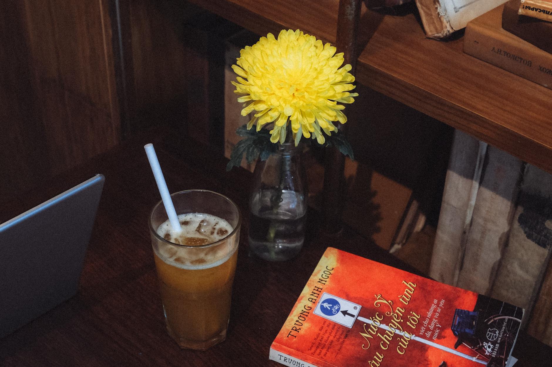 A warm café table with a yellow chrysanthemum, iced coffee, and a book in Ho Chi Minh City, Vietnam.
