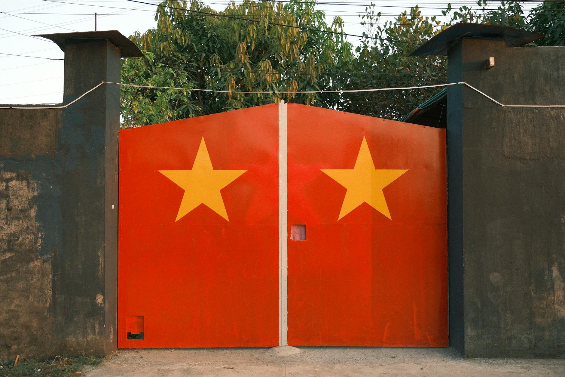 Red metal gate with two yellow stars, outdoors with green foliage in the background.