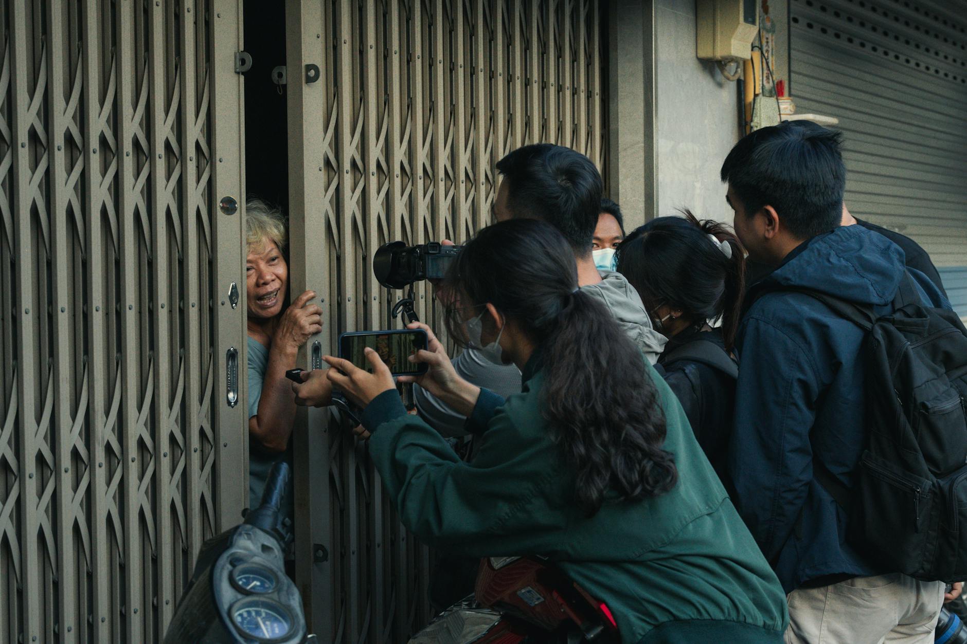 A group of people engage with an elderly woman behind a metal gate on a city street.