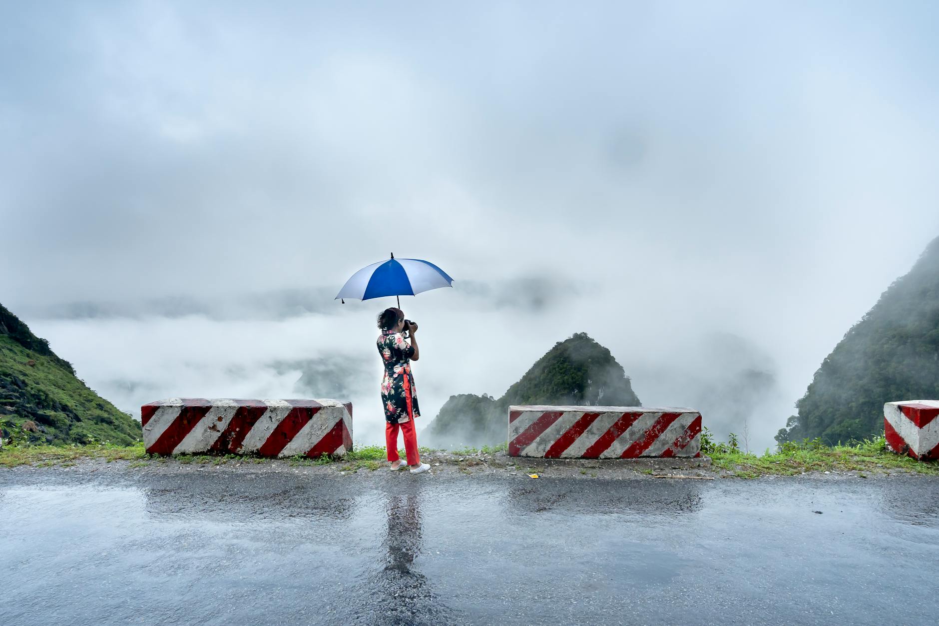 A woman stands with an umbrella on a misty mountain road, overlooking fog-covered hills.