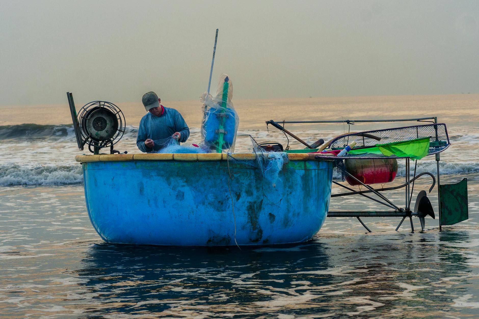 A Vietnamese fisherman in a traditional blue basket boat prepares fishing nets by the shore.