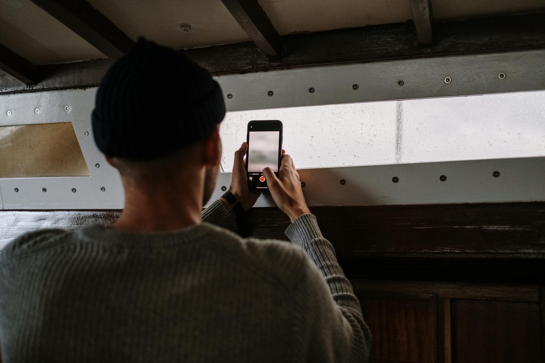 A man in a gray sweater takes a picture with his smartphone inside a wooden room.