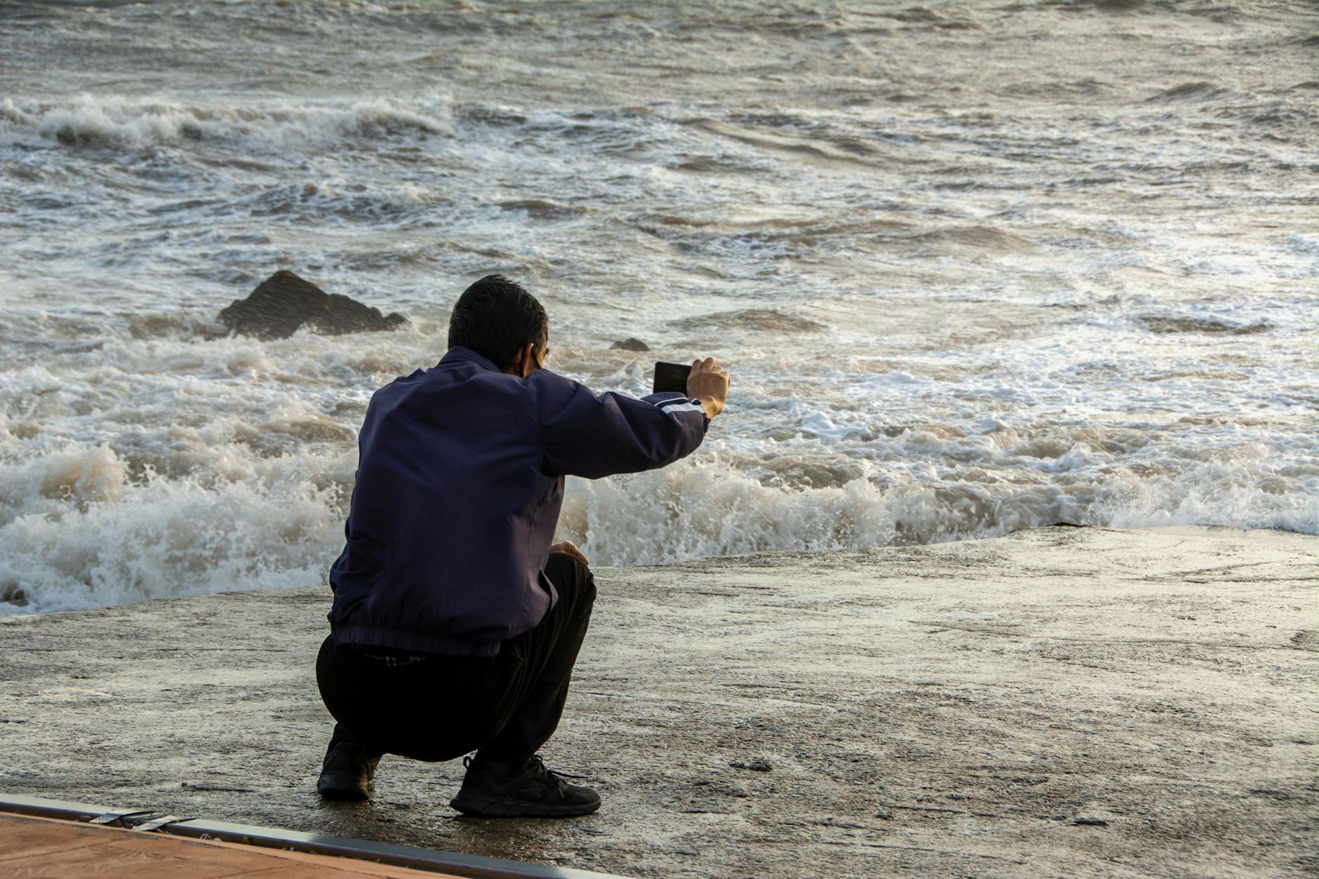 A man squats by the shore, capturing the crashing ocean waves with his smartphone.
