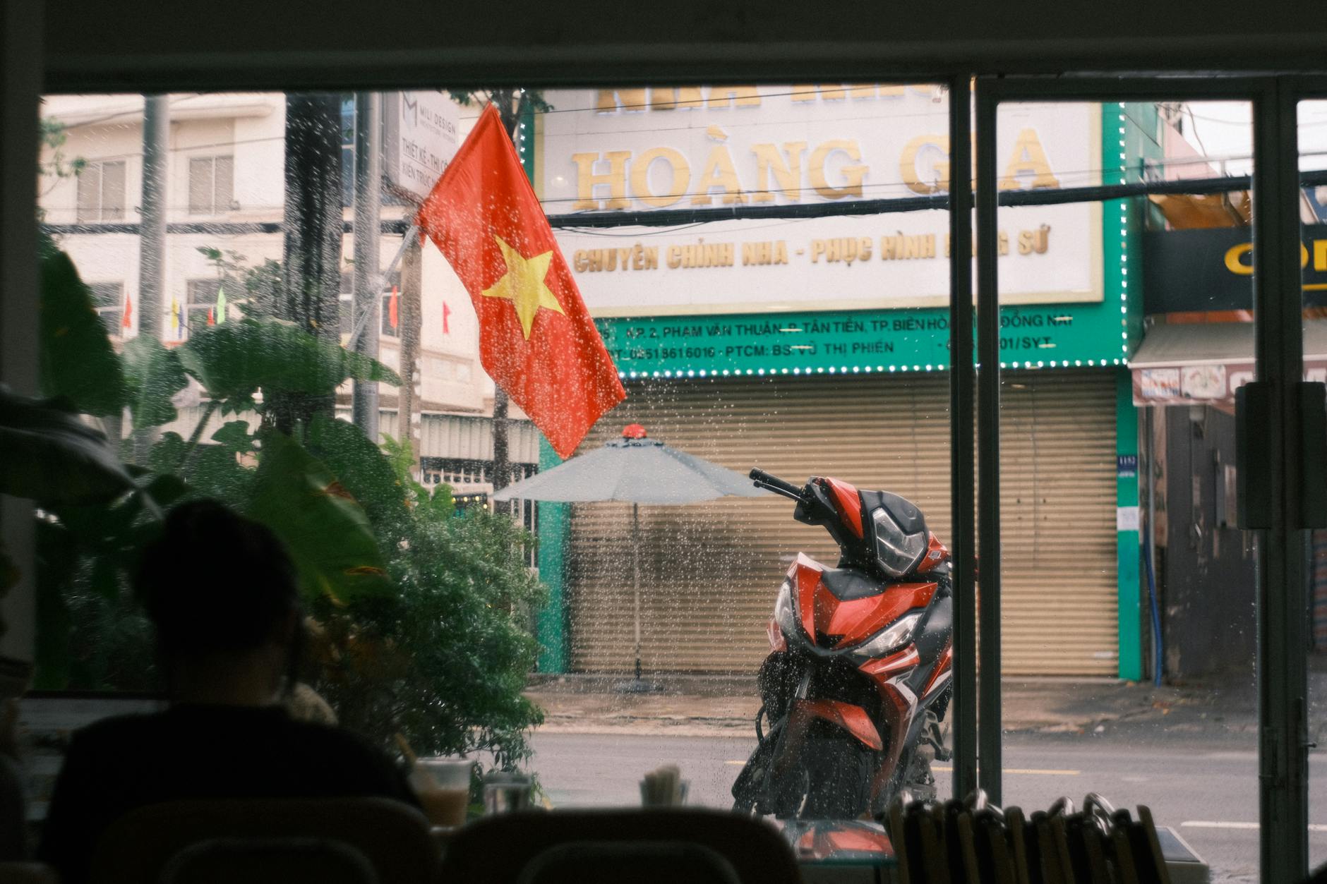 A Vietnamese street with a flag and scooter seen through a cafe window during rain.