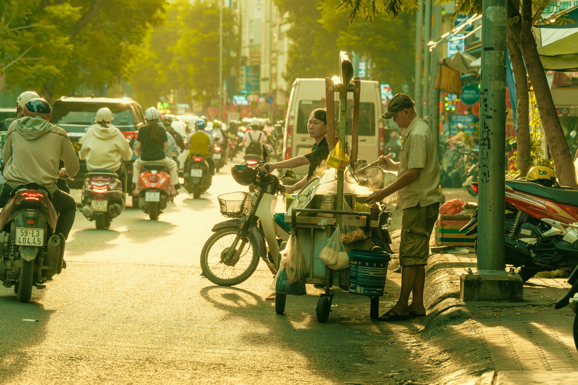 Crowded city street with people on motorcycles and a street food vendor serving customers under warm sunlight.