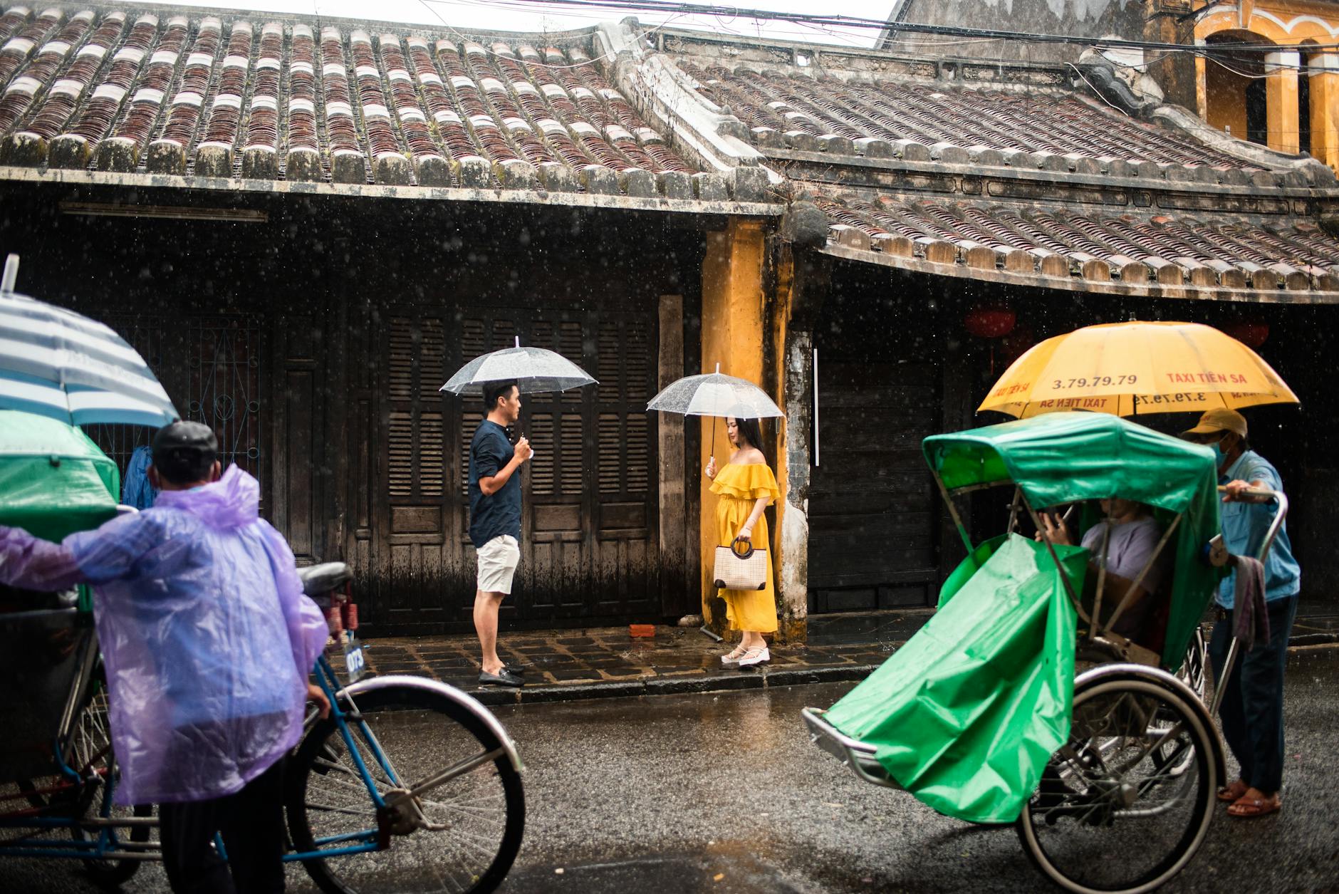 People walking on a rainy street with umbrellas, alongside bicycles and a traditional building facade.