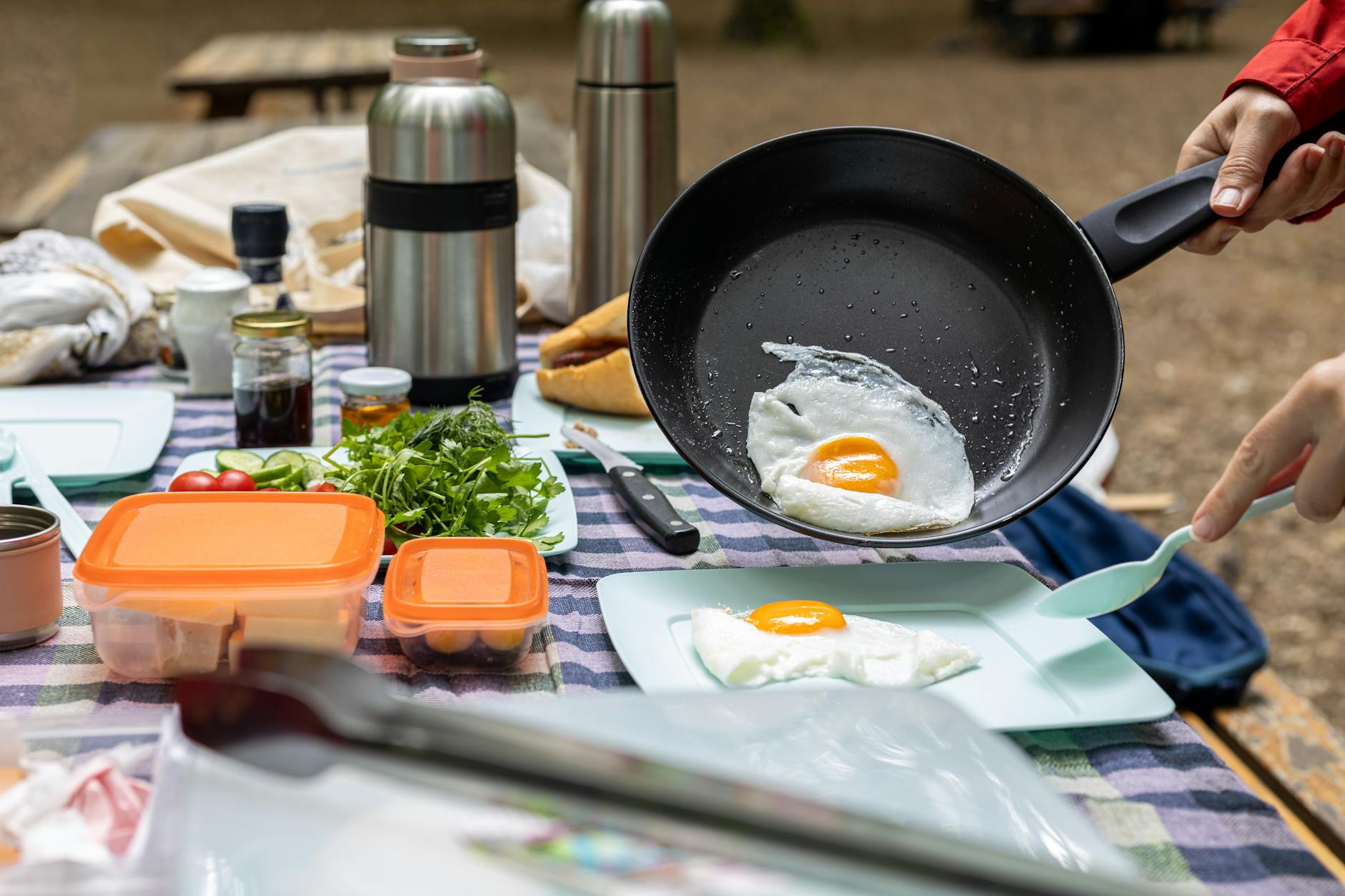 Campers cooking fried eggs outdoors with fresh greens, tomatoes, and thermos.