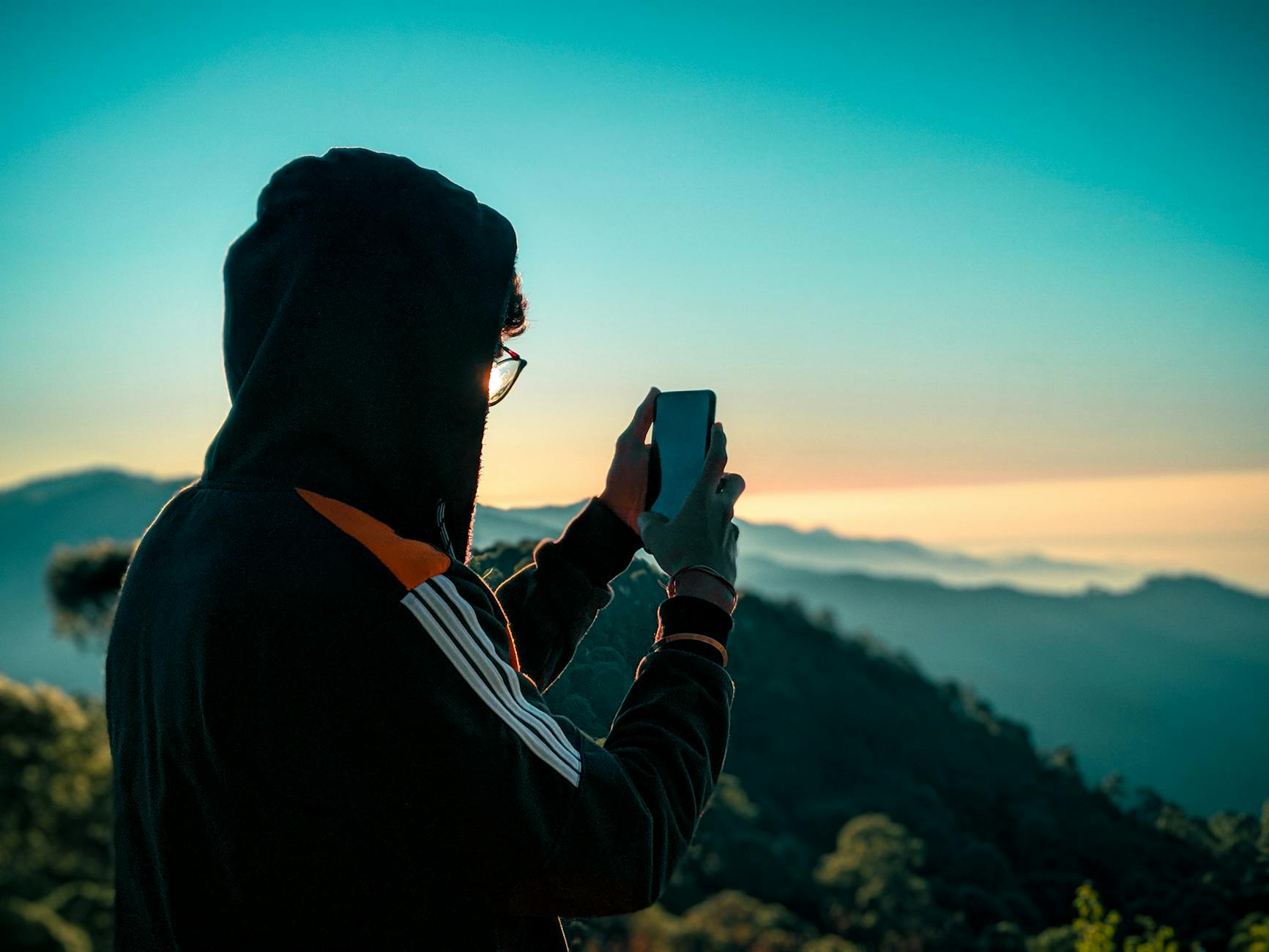 Person in a hoodie taking a photo of a scenic mountain view on a smartphone during the day.