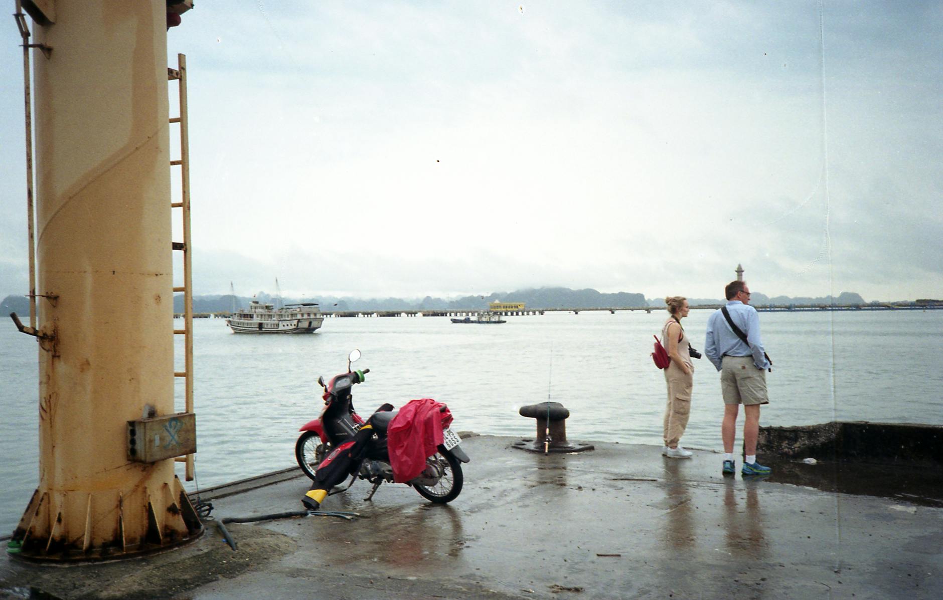 Couple stands on a pier at Ha Long Bay, Vietnam, with a ship in the distance.