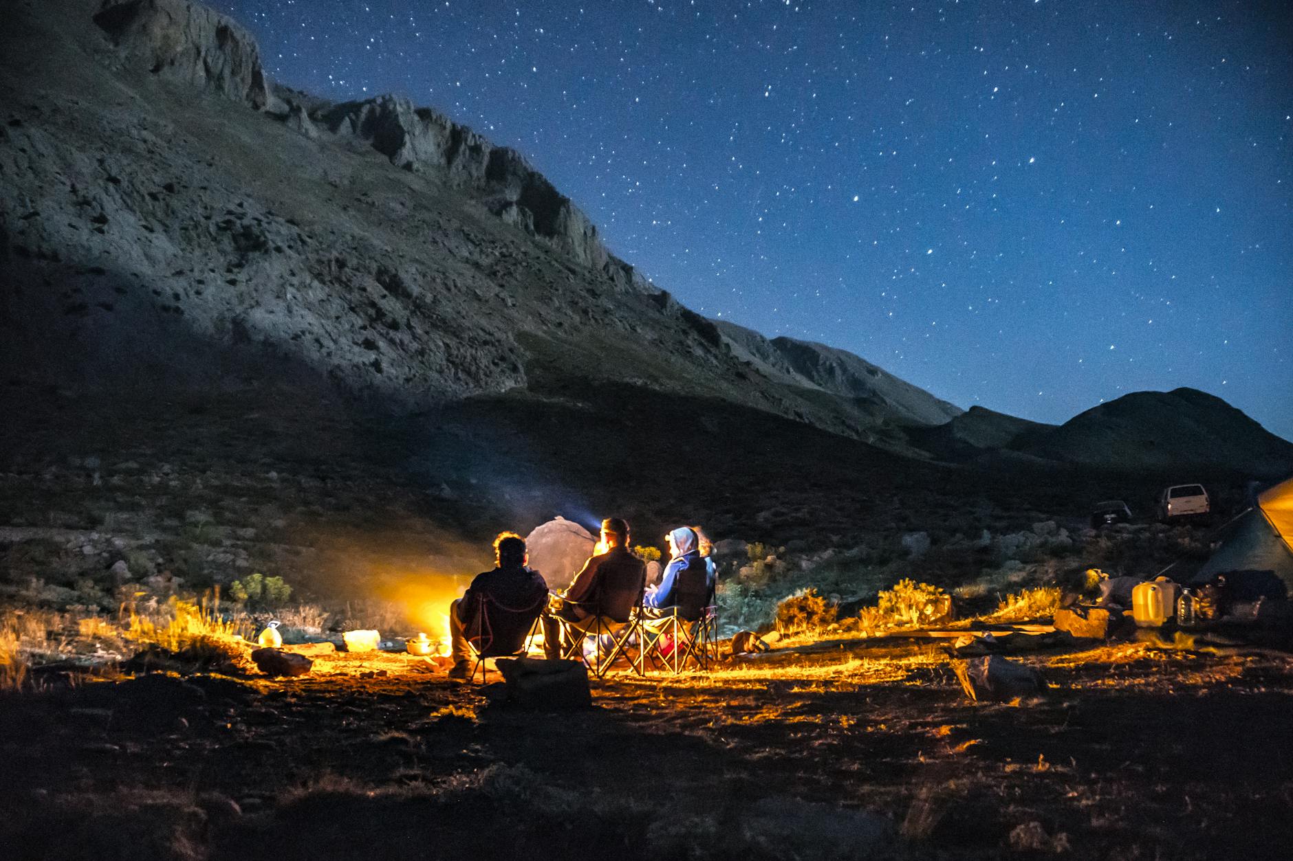 Group of adults enjoying a campfire under a starry sky in the mountains.
