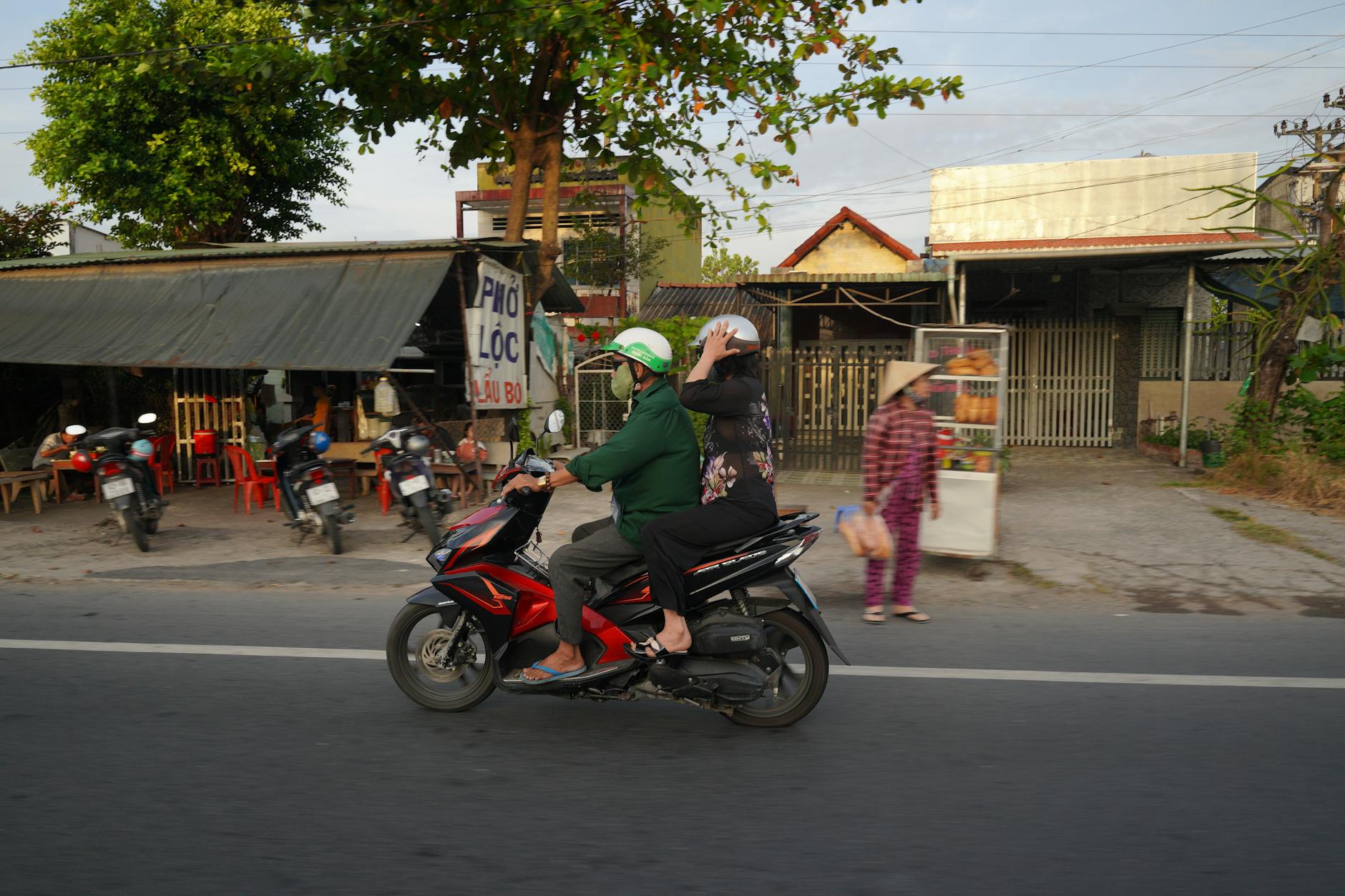 A motorbike with two riders passes by roadside shops on a busy urban street.
