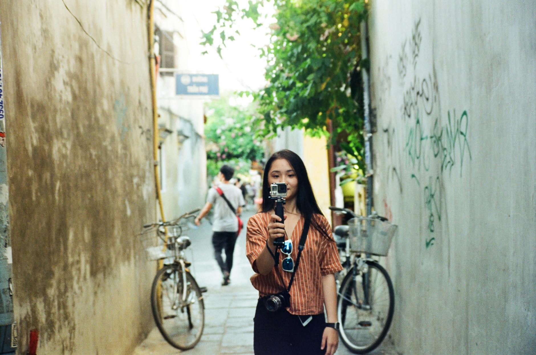 A young woman holds a camera while walking through a narrow urban alley with bicycles in the background.
