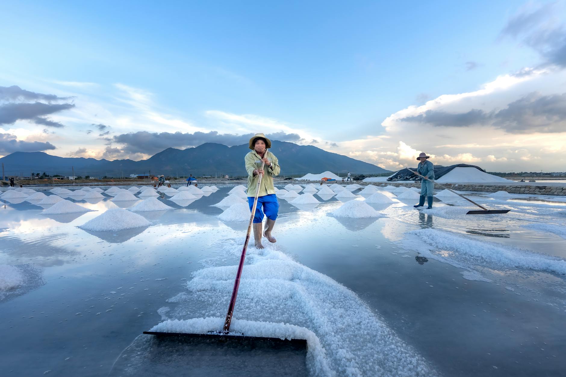 Workers gather salt at a serene salt farm, under a vibrant morning sky.
