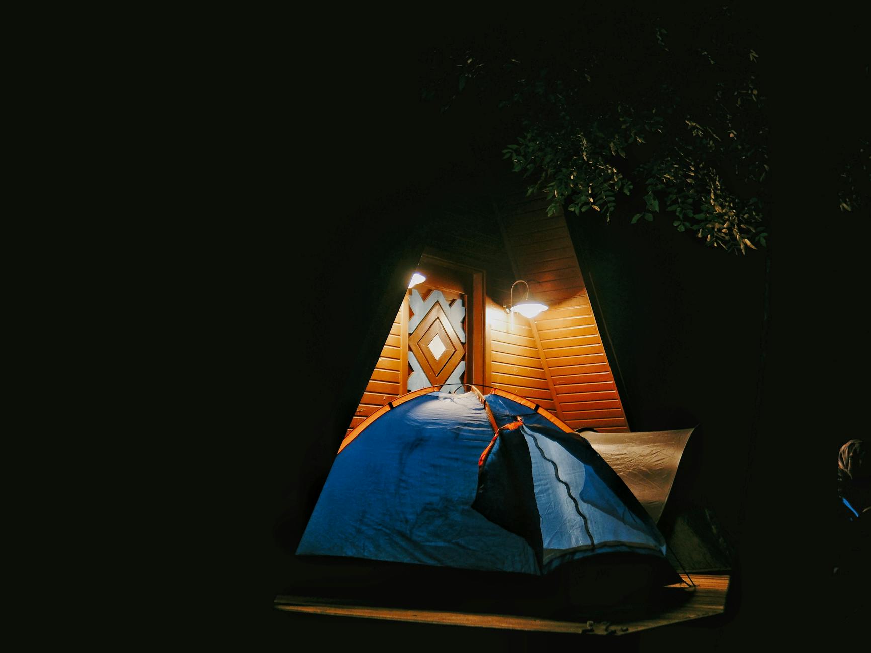 A cozy blue tent illuminated at night beside a rustic A-frame cabin under a tree.