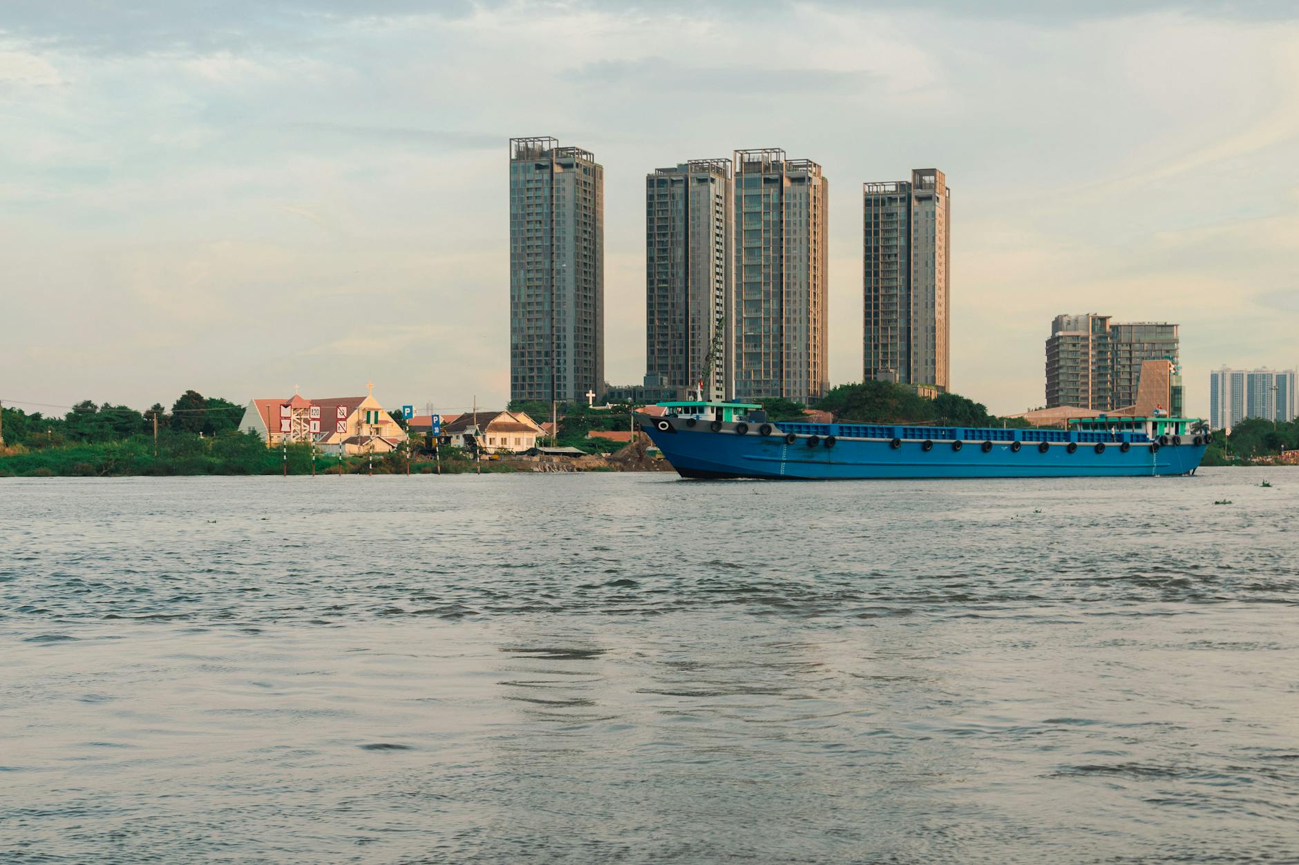 A serene view of a blue cargo ship passing city buildings on a calm river at sunset.