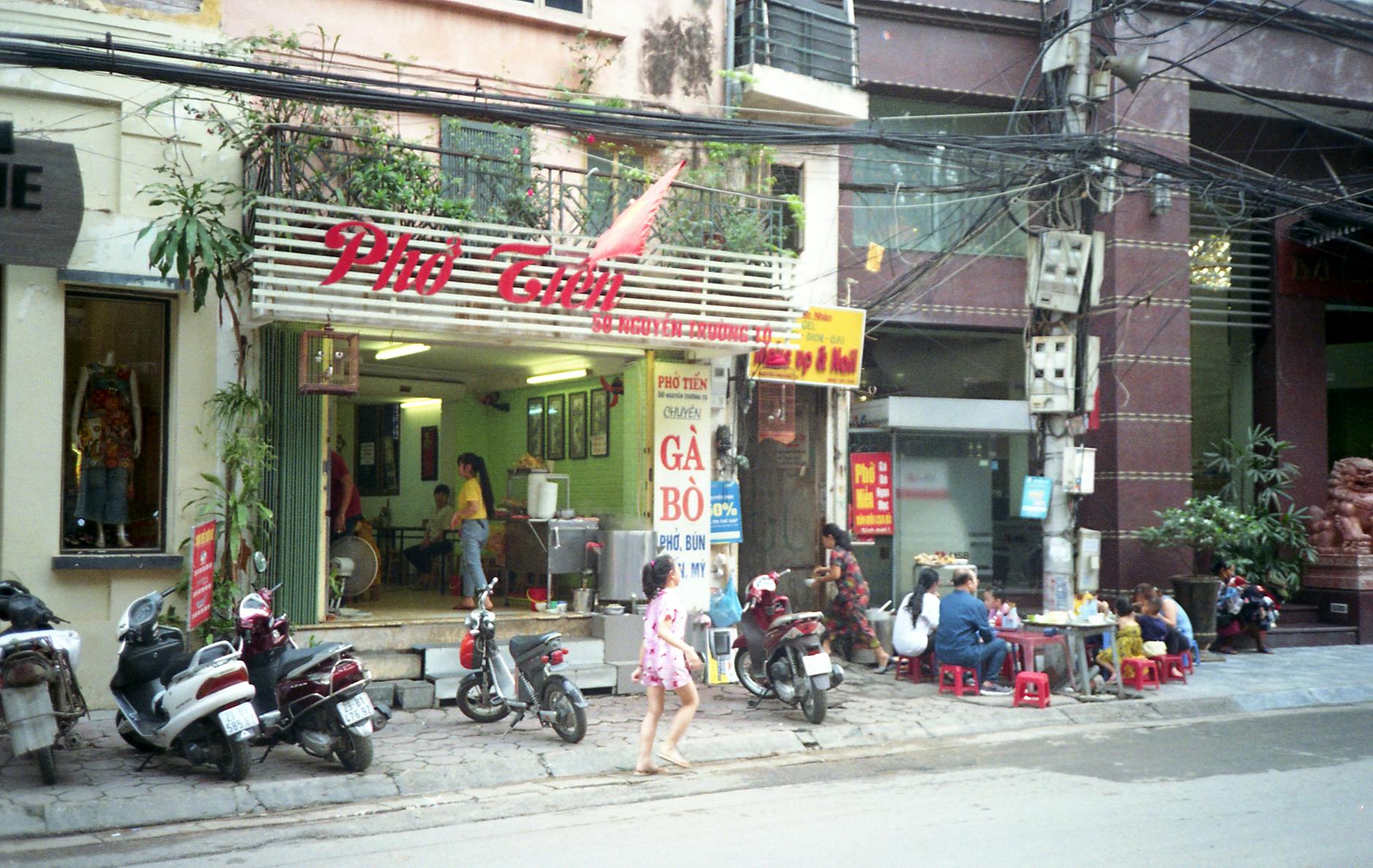 An outdoor scene of a bustling Vietnamese restaurant with people dining on the sidewalk in Hanoi.