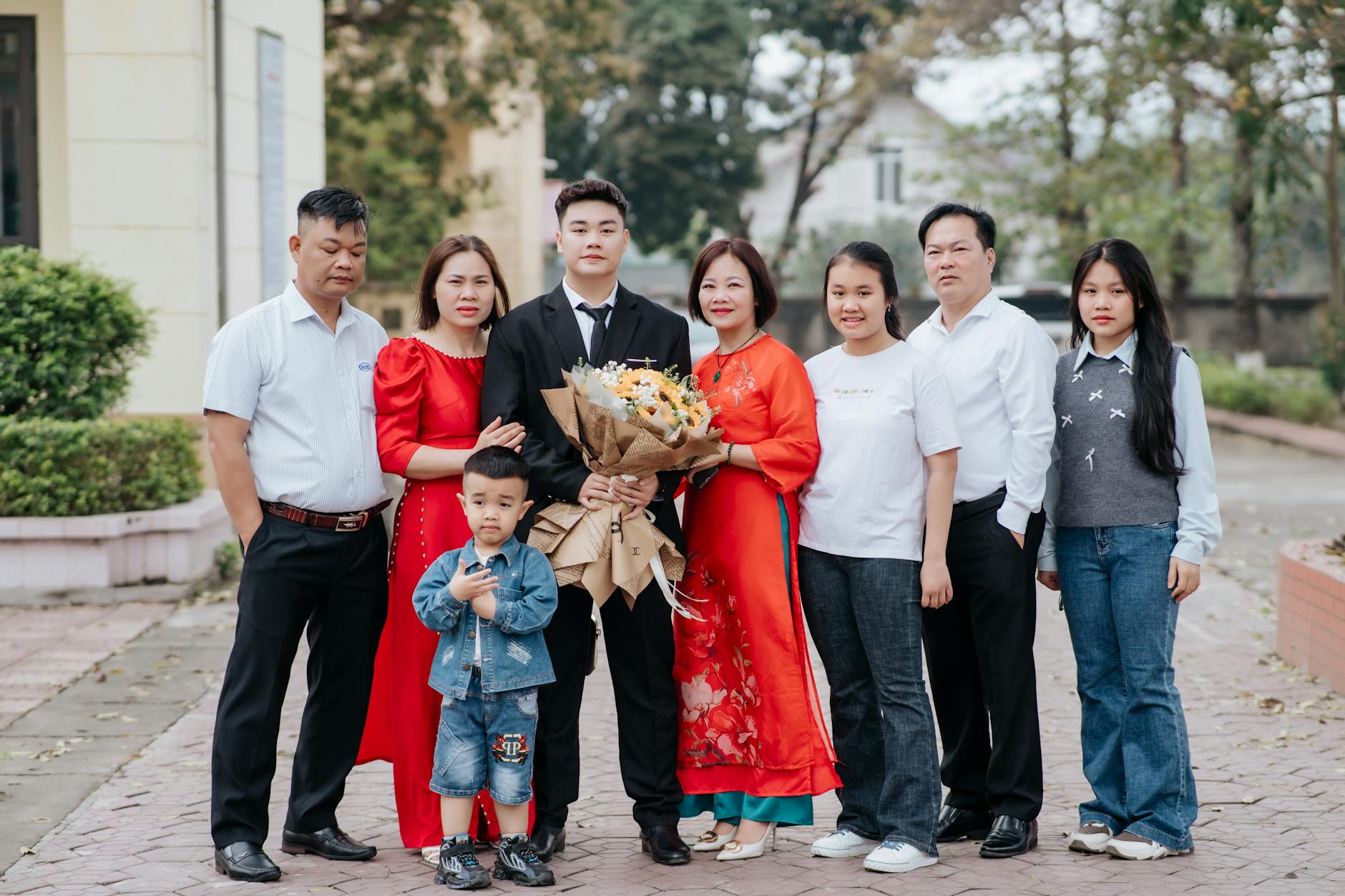 Group family portrait outdoors with a formal dressed individual holding flowers, surrounded by relatives.