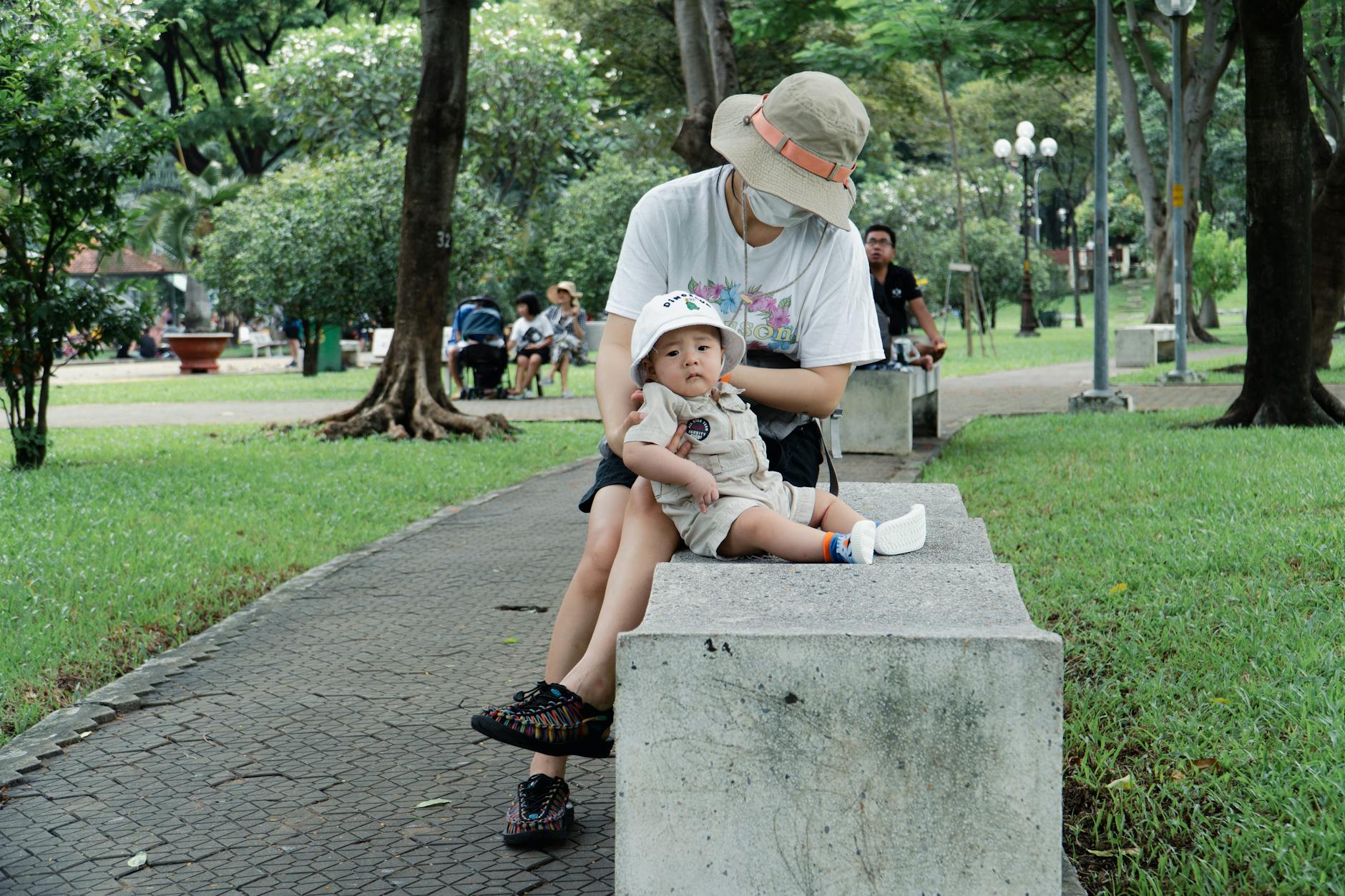A serene moment of a mother and child seated on a bench in a lush Ho Chi Minh park during a summer day.