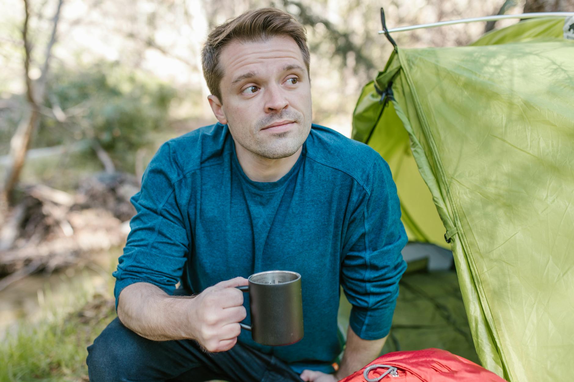 Man enjoying morning coffee by a tent in a lush outdoor setting.