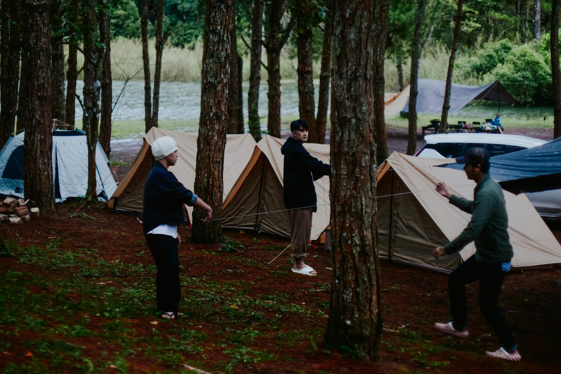 Three friends setting up tents in a lush forest campsite by the water.