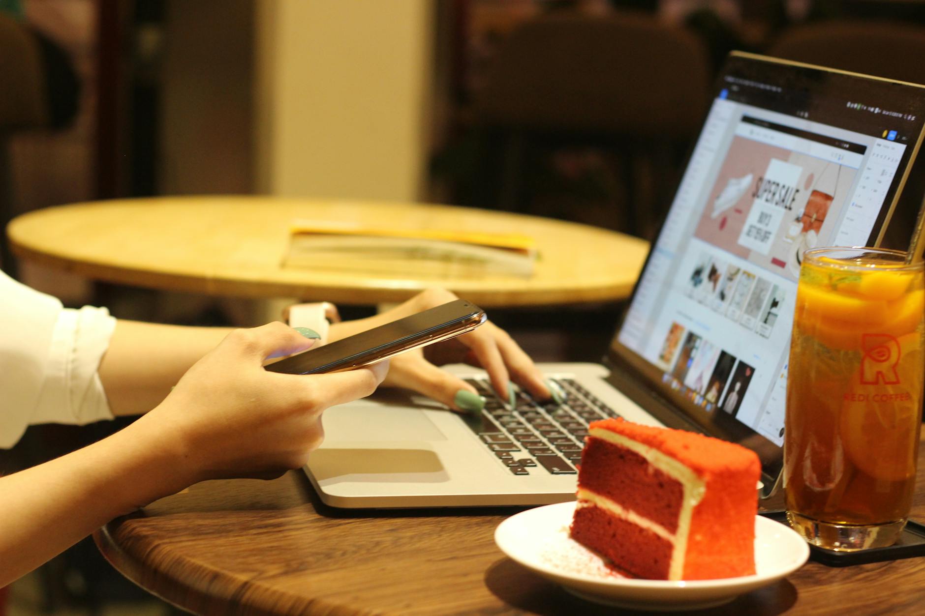 A woman works on a laptop while using her smartphone in a cozy coffee shop.