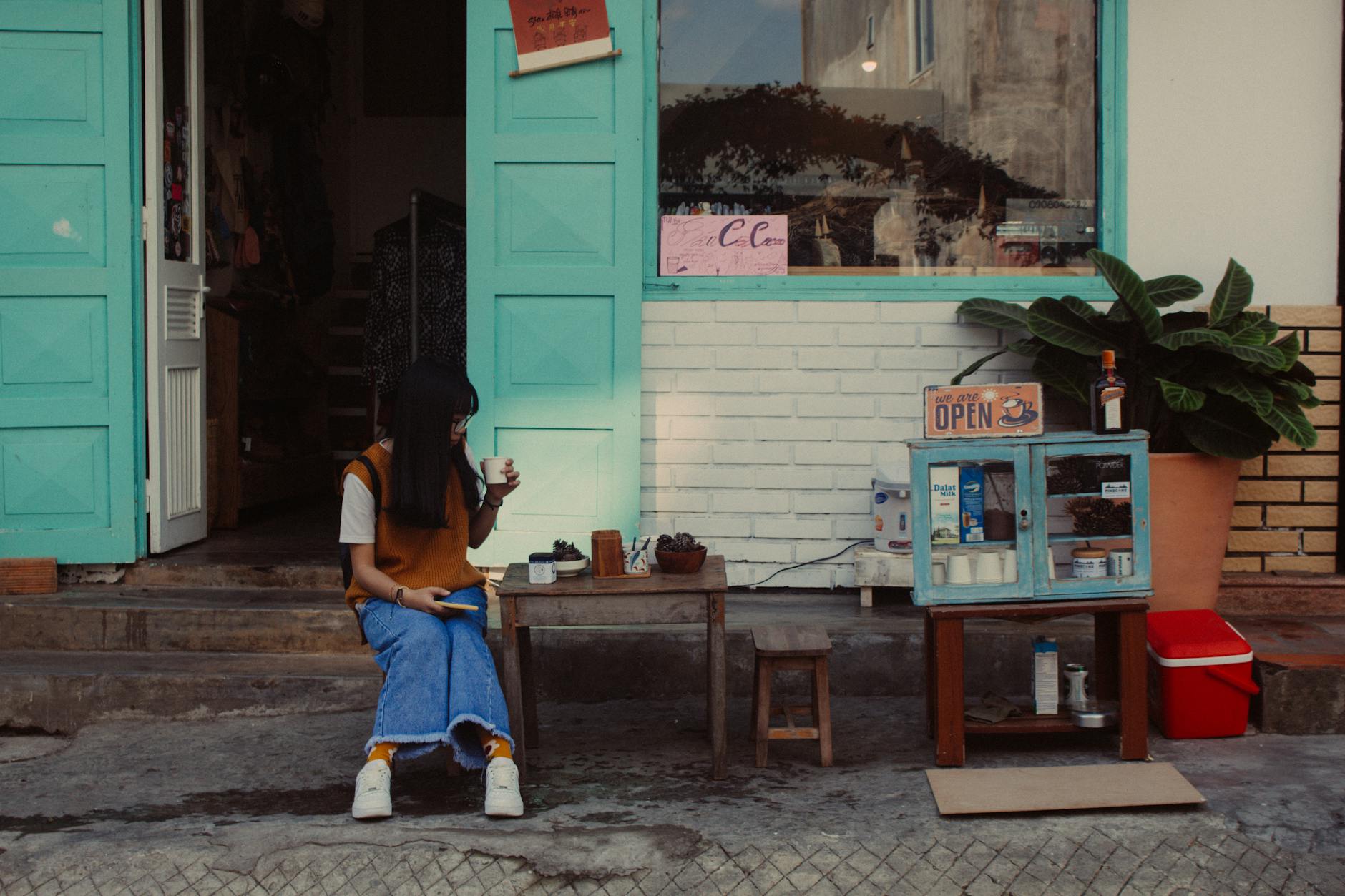 Young woman enjoys coffee outdoors at a charming street shop with vintage decor.