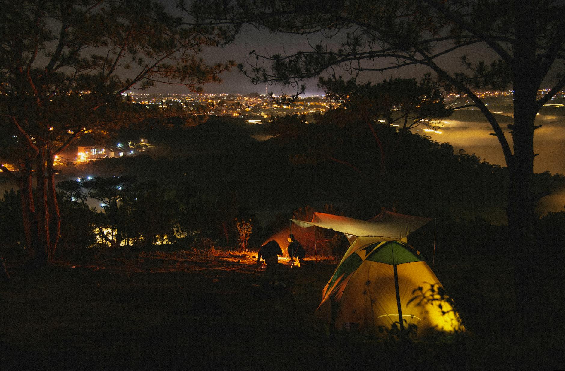 A serene camping scene at night with tents and a bonfire under a night sky and city lights in the distance.