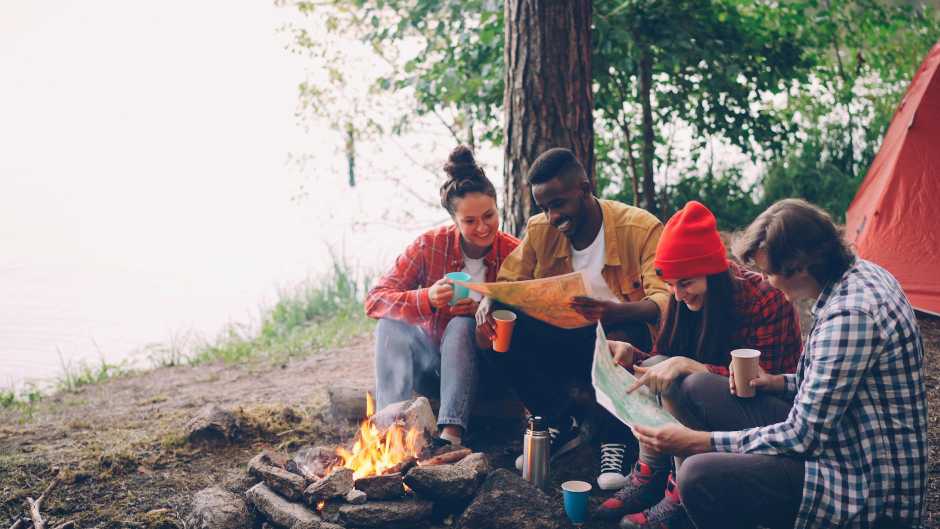 Group of friends camping outdoors, enjoying a campfire by the lakeside with maps.