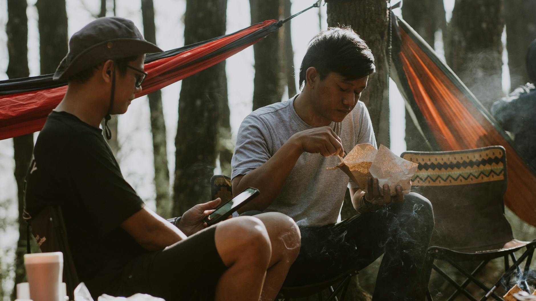 Two men enjoy a casual meal and smartphone browsing in a forest hammock setup.