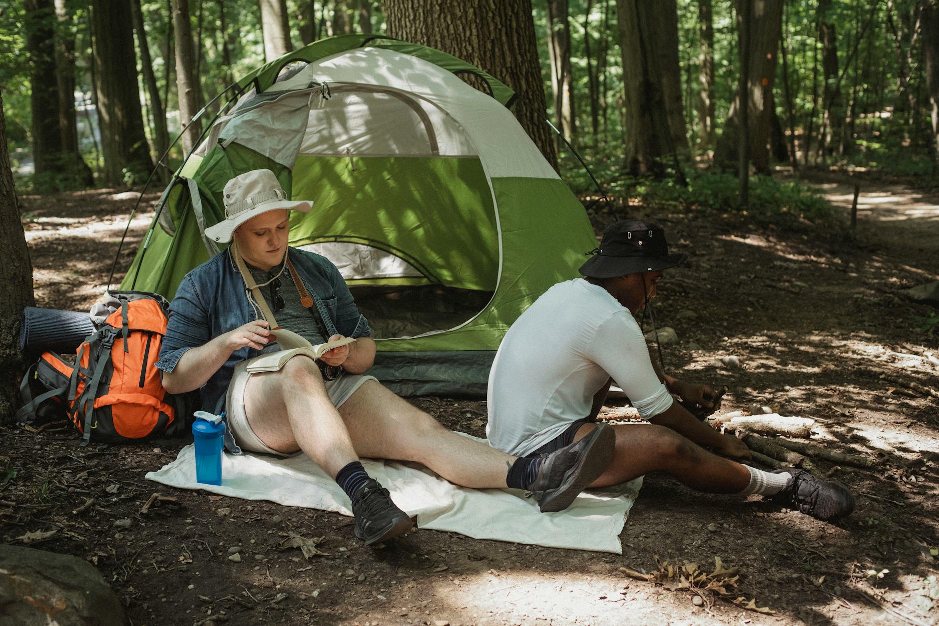 Concentrated young guy sitting on blanket and reading book while camping together with black friend in summer woods
